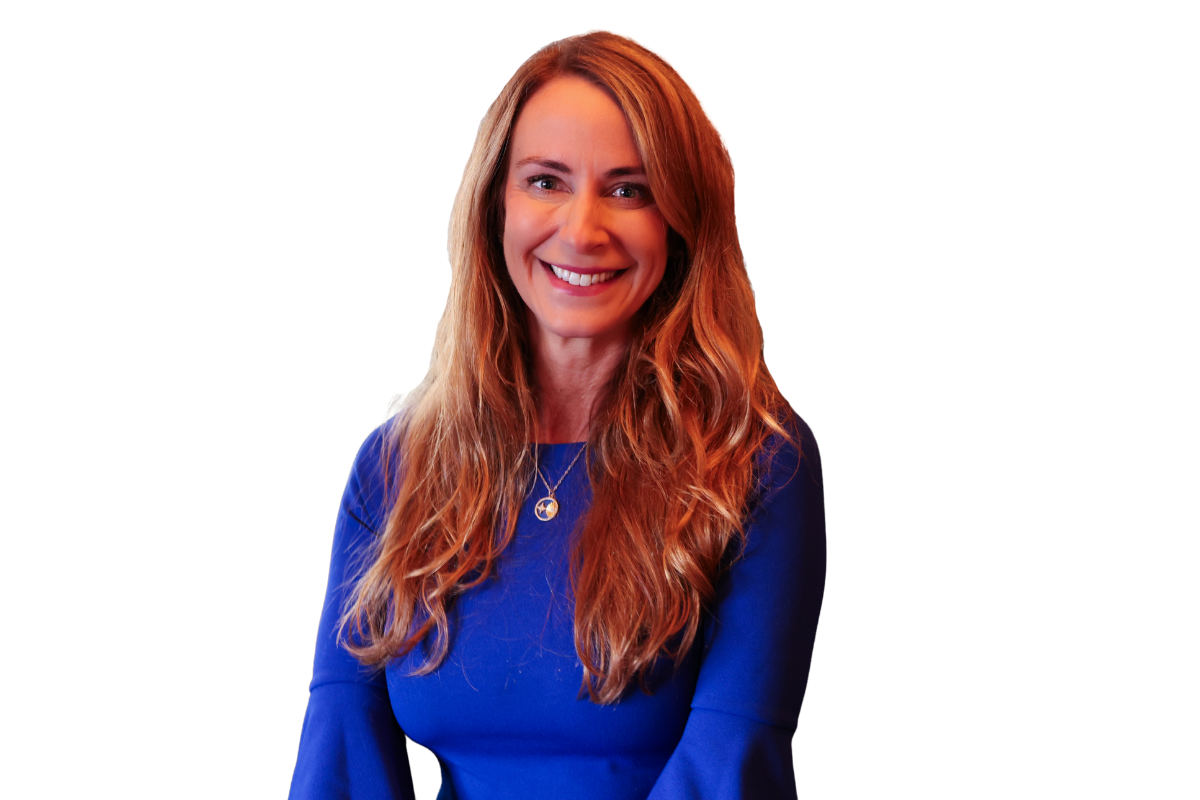 A woman with long red hair smiling, wearing a blue top and a peace sign necklace.