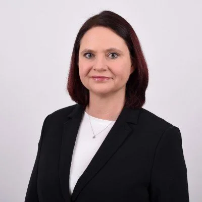 A woman with shoulder-length dark red hair, wearing a black blazer over a white top, standing against a plain white background.
