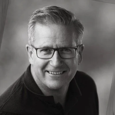 Black and white photo of a smiling man with glasses and short, neatly styled hair, wearing a dark shirt.