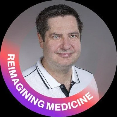 A man with dark hair and light skin, wearing a white polo shirt with black trim, smiling at the camera, with a circular background that says 'REIMAGINING MEDICINE' in white and purple text.