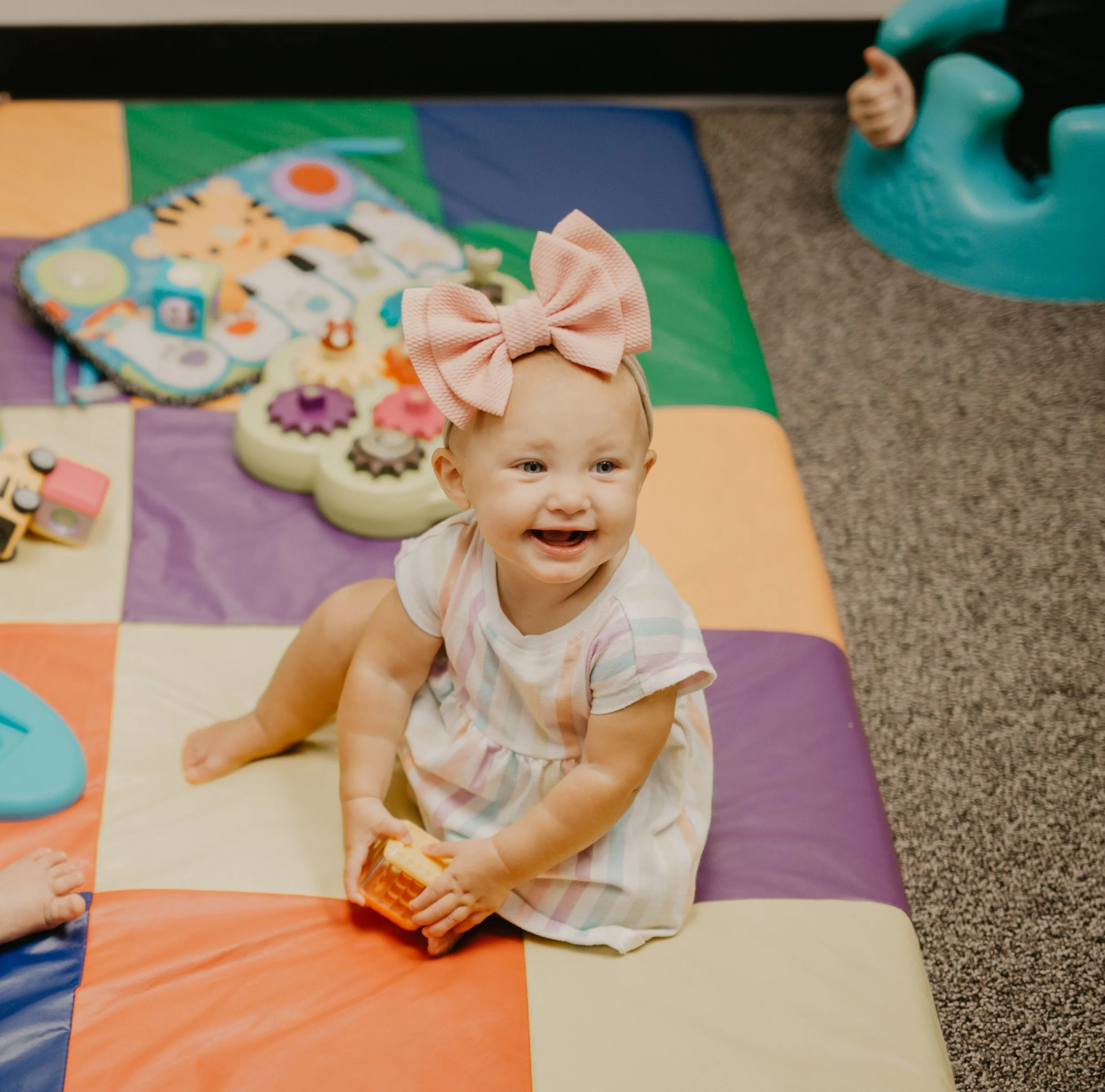 A smiling baby girl with a large pink bow headband, wearing a pastel striped dress, sitting on a multicolored foam play mat, holding a small toy block, with toys and a blue baby chair nearby.