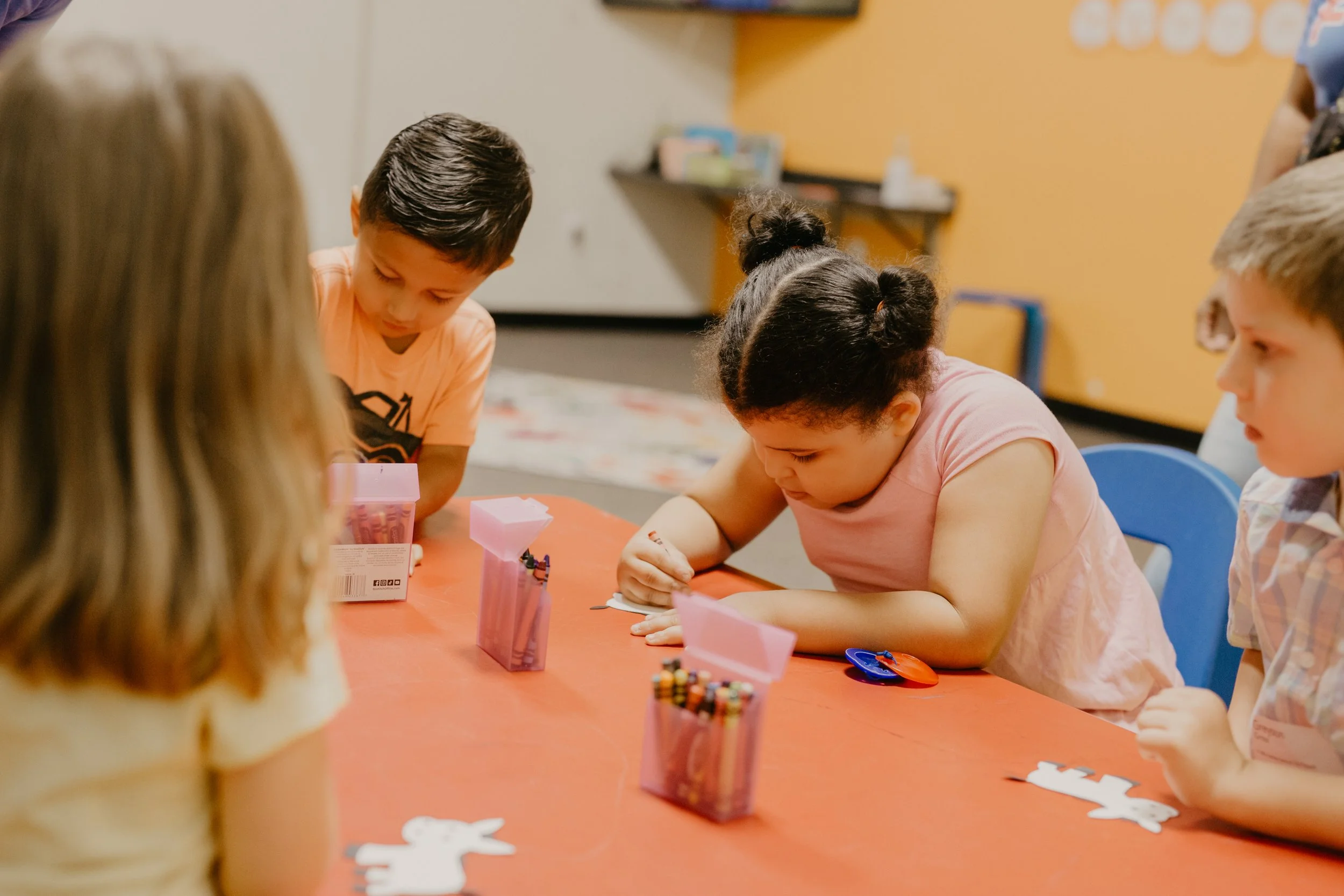 Children working on a craft project at a table with pink containers of art supplies in a classroom.
