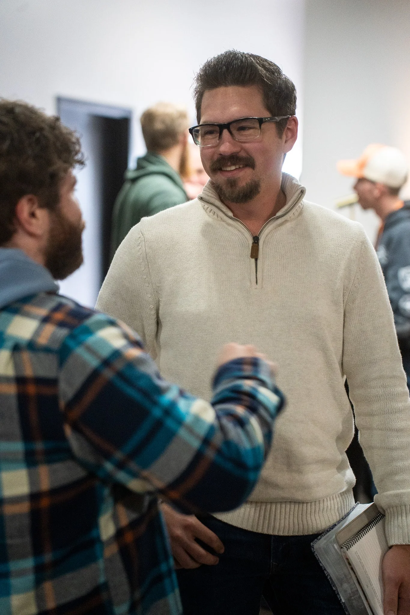 Two men engaged in conversation at an indoor gathering, with other people in the background.