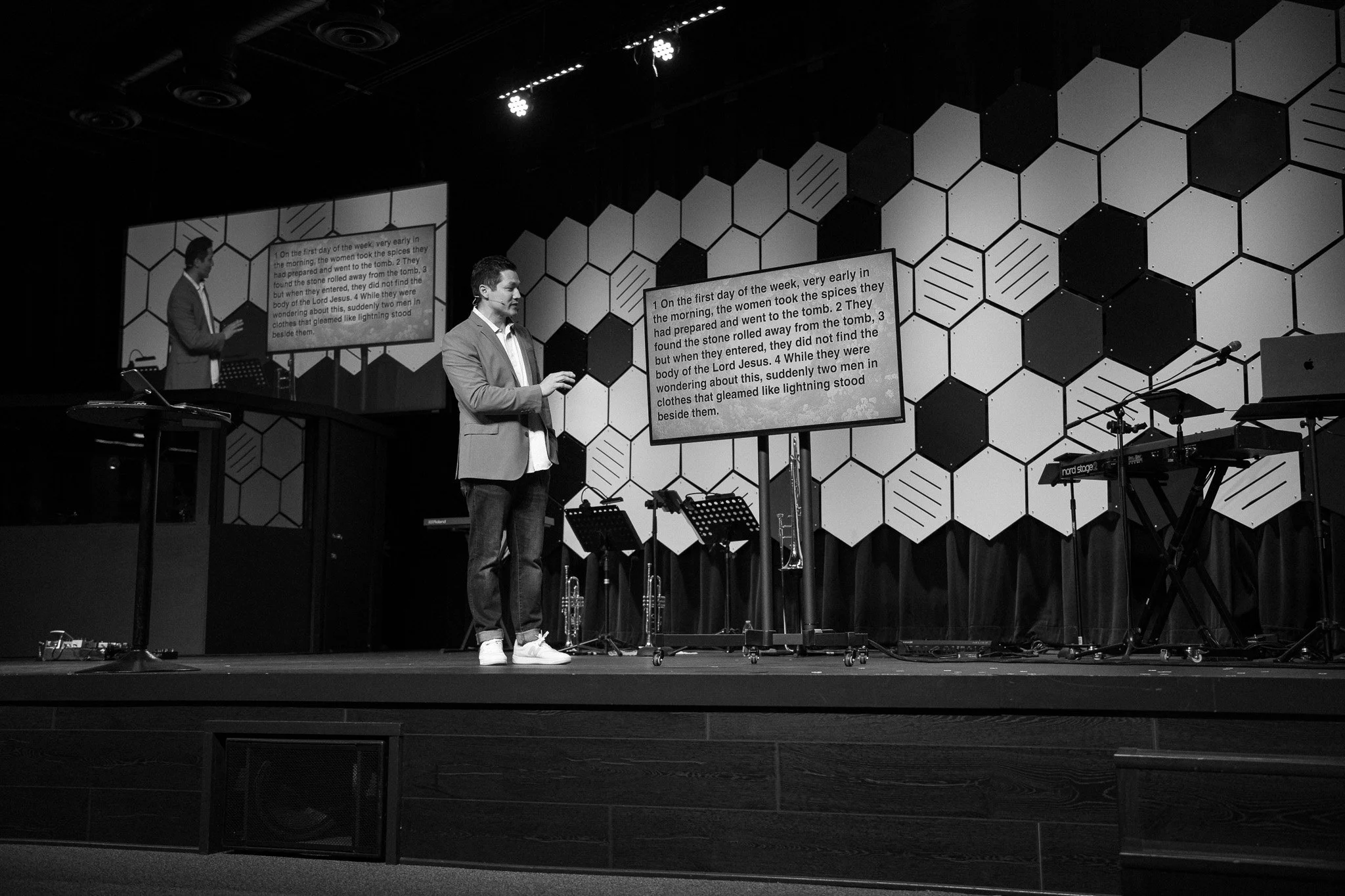 A man on stage giving a presentation with a large screen displaying text behind him, featuring musical instruments and a geometric patterned wall.