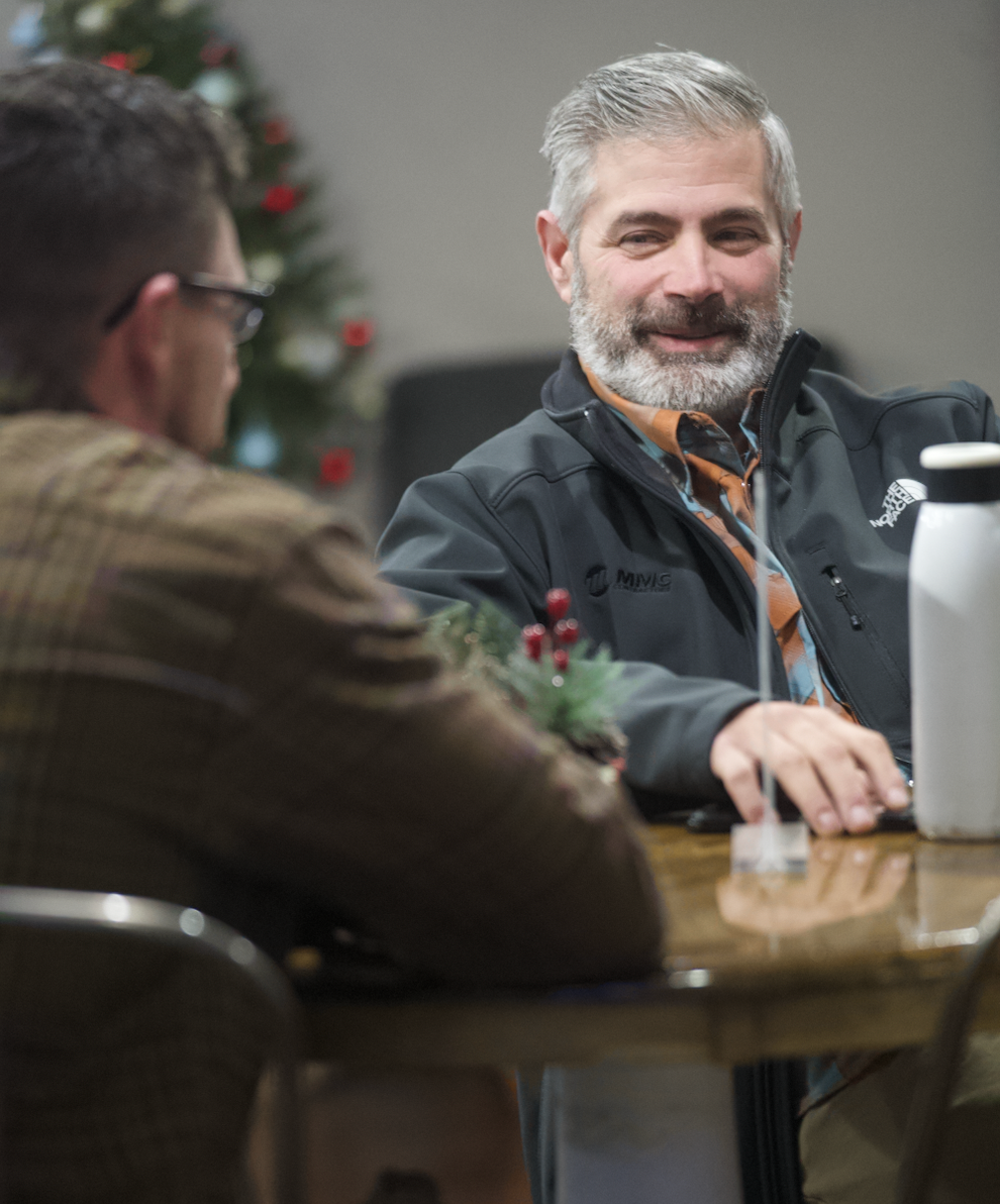 Two men sitting at a table, smiling and talking, with a Christmas tree in the background. One man has gray hair and a beard, wearing a black jacket, and the other man has dark hair, wearing glasses and a brown plaid shirt.