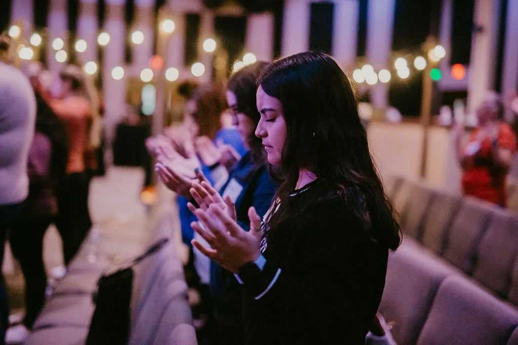 Women praying with eyes closed at a gathering with string lights overhead.