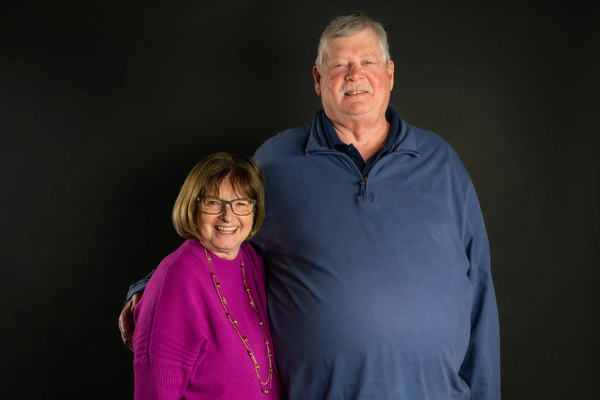 A smiling woman with glasses and brown hair wearing a bright pink sweater standing next to a tall man with gray hair wearing a blue long-sleeve shirt against a black background.