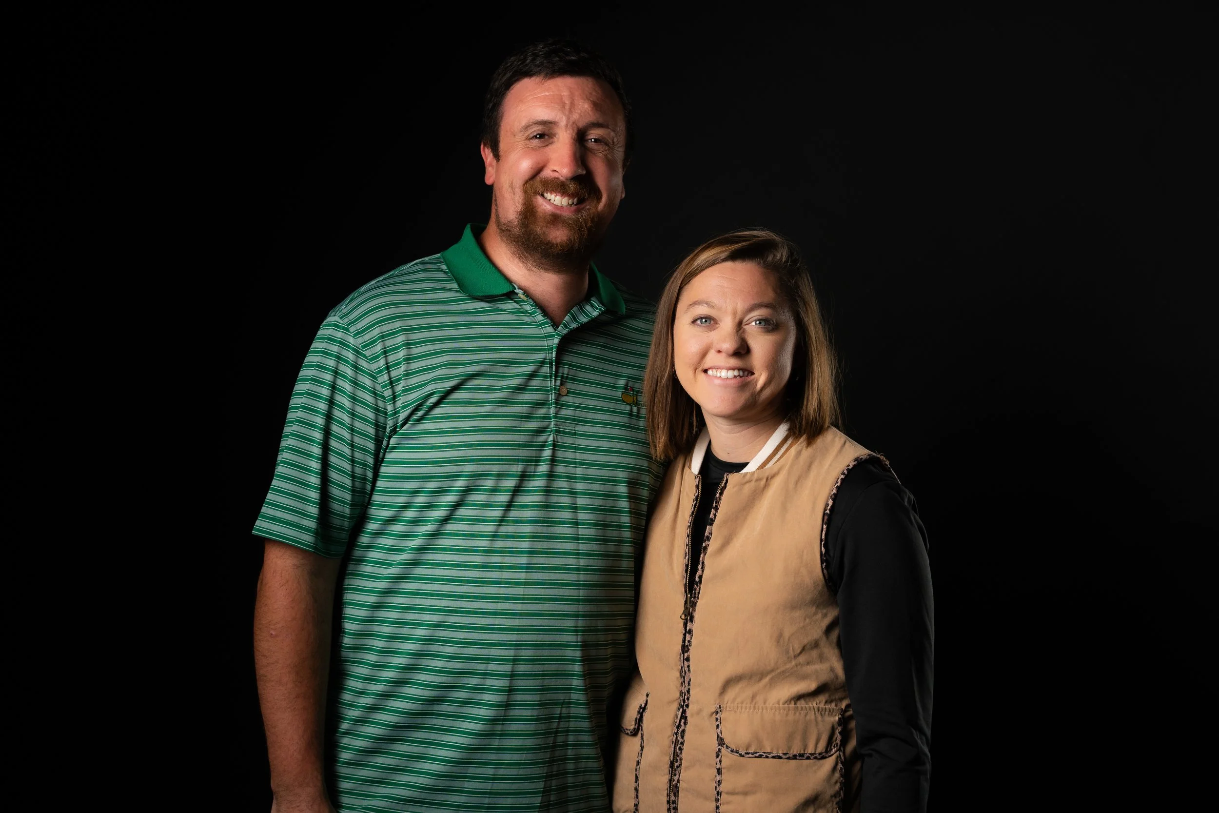 A smiling man with a beard wearing a green striped polo shirt stands beside a smiling woman with shoulder-length brown hair wearing a black shirt and a beige vest, both posed against a black background.