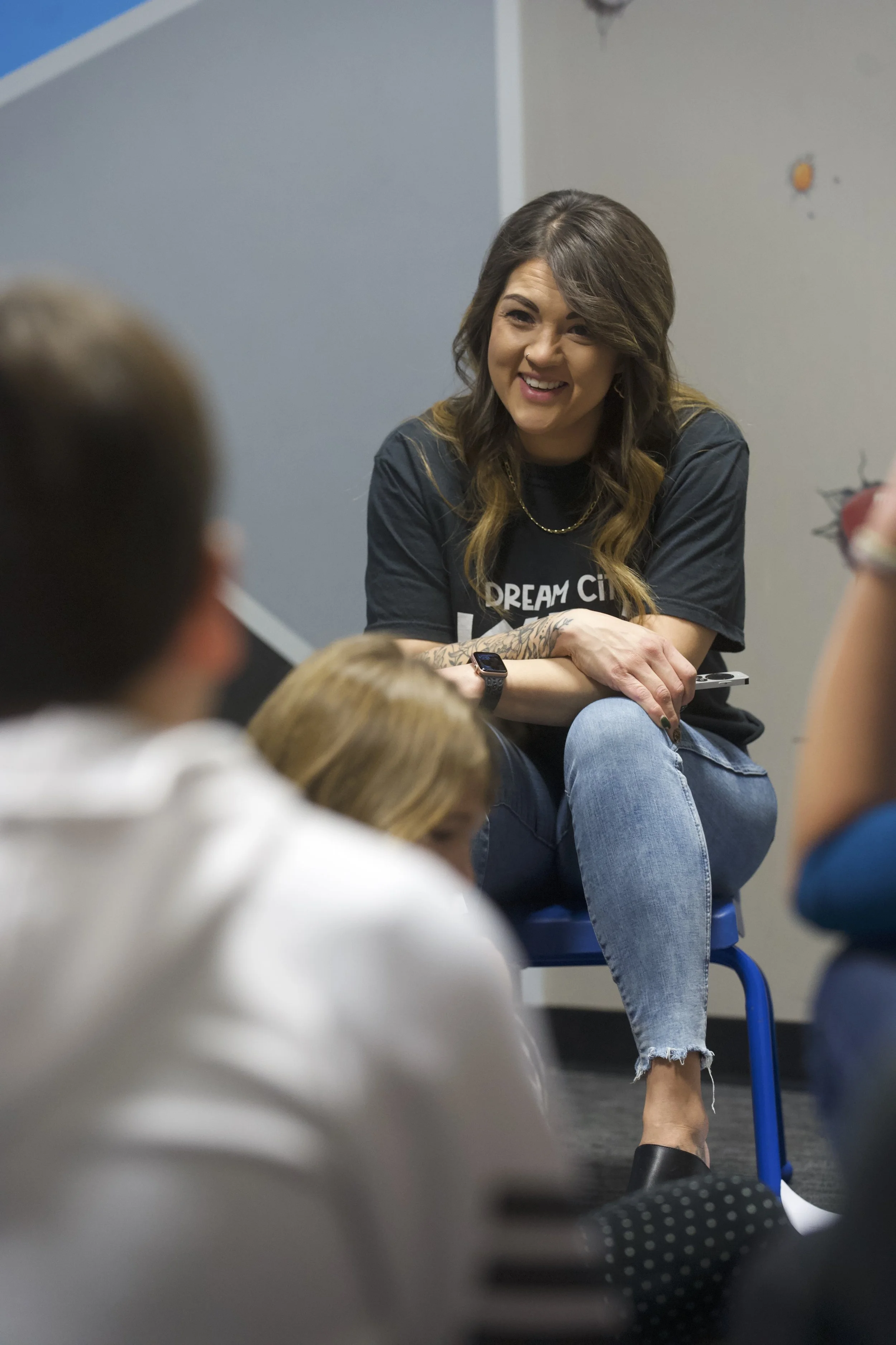 A woman with long hair, wearing a Dream City t-shirt, sitting on a chair, engaging with a group of children sitting on the floor, in a classroom setting.