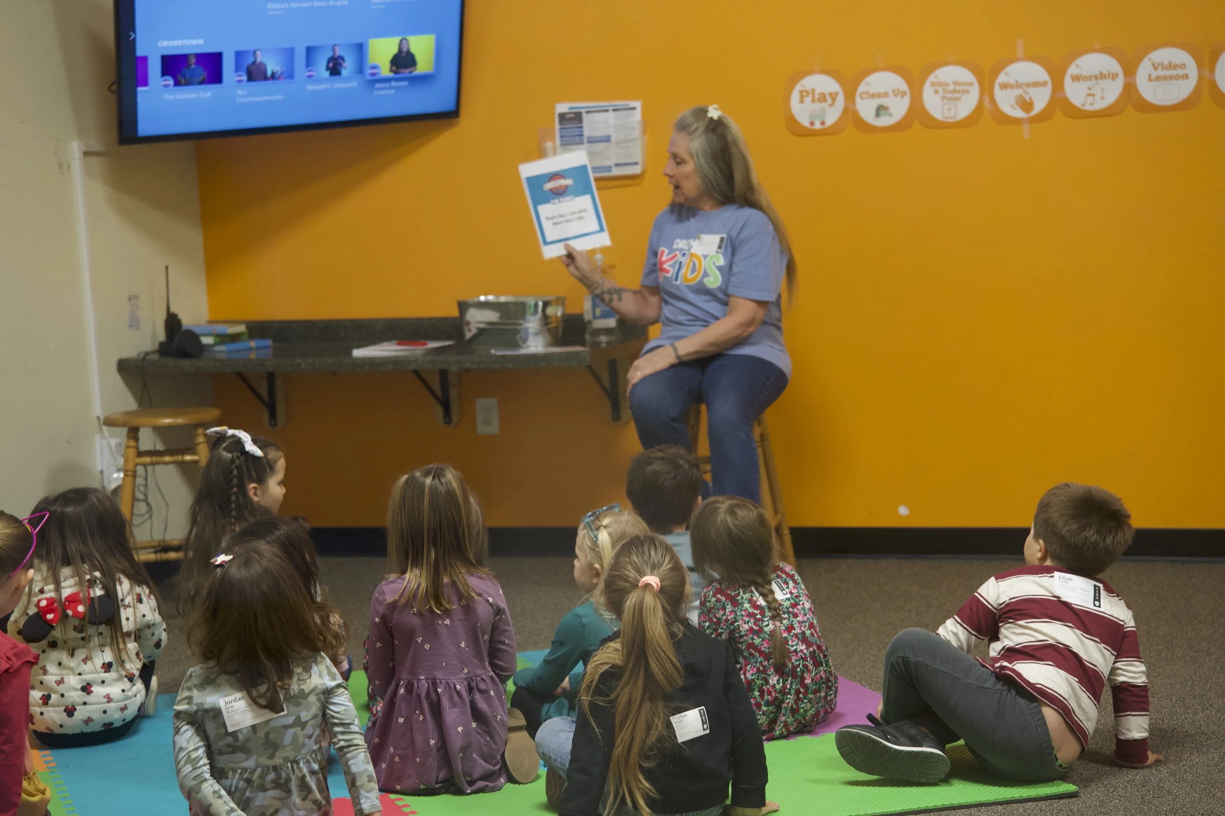 A woman sitting on a stool reading to a group of children seated on colorful mats in a classroom with orange walls. There is a large screen on the wall displaying a television show, and a table behind the woman with books and papers.