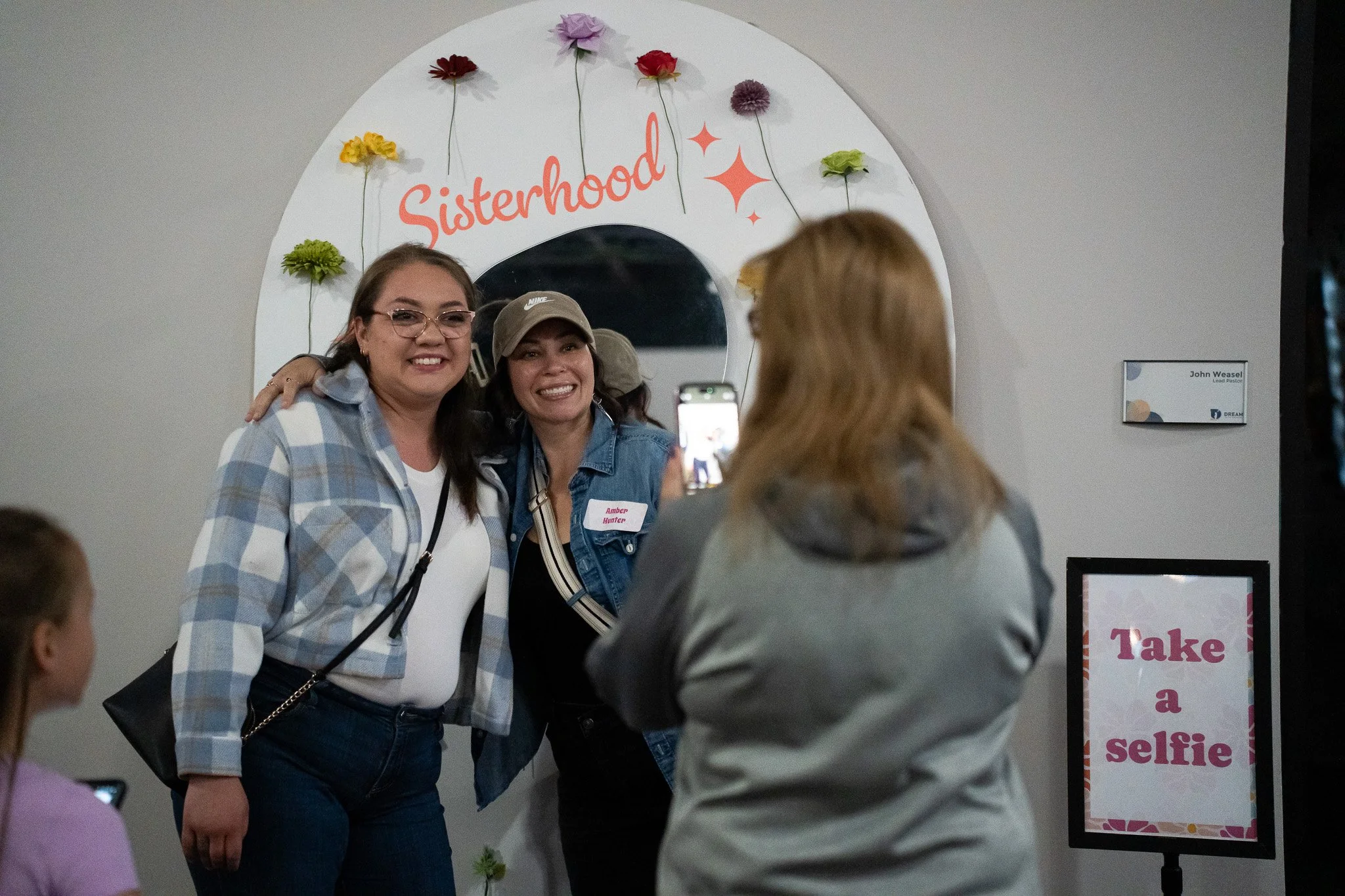 Two women are posing for a selfie in front of a decorative wall with flowers and the word "Sisterhood" on it. One woman is wearing glasses, a plaid shirt, and jeans, while the other is wearing a denim jacket and a baseball cap. A person with long hair and a gray top is taking their photo. There are also two other women, one on the left side and one partially visible in the foreground.