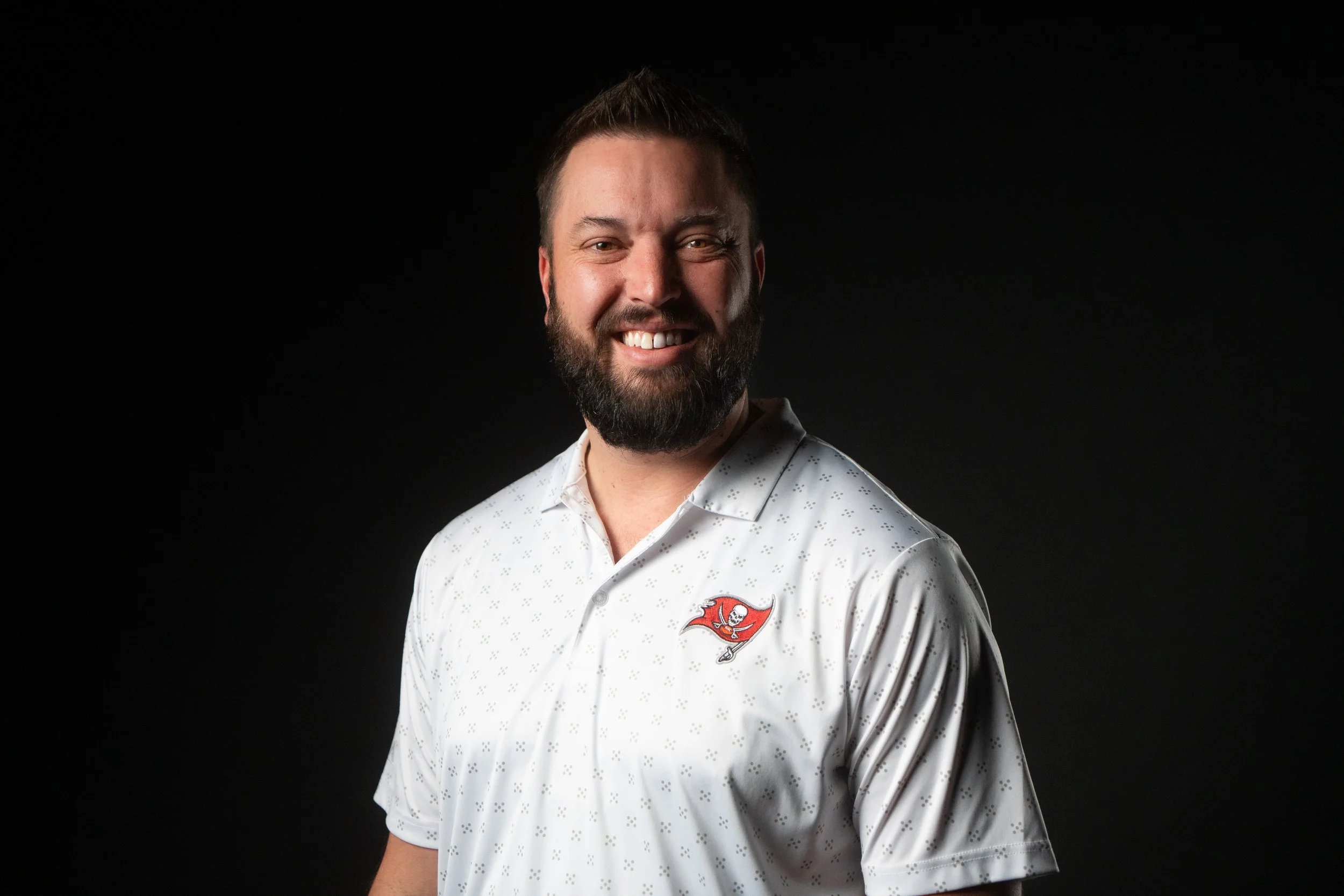 A smiling man with a beard and short hair wearing a white Tampa Bay Buccaneers polo shirt with a small Buccaneers logo, standing against a black background.