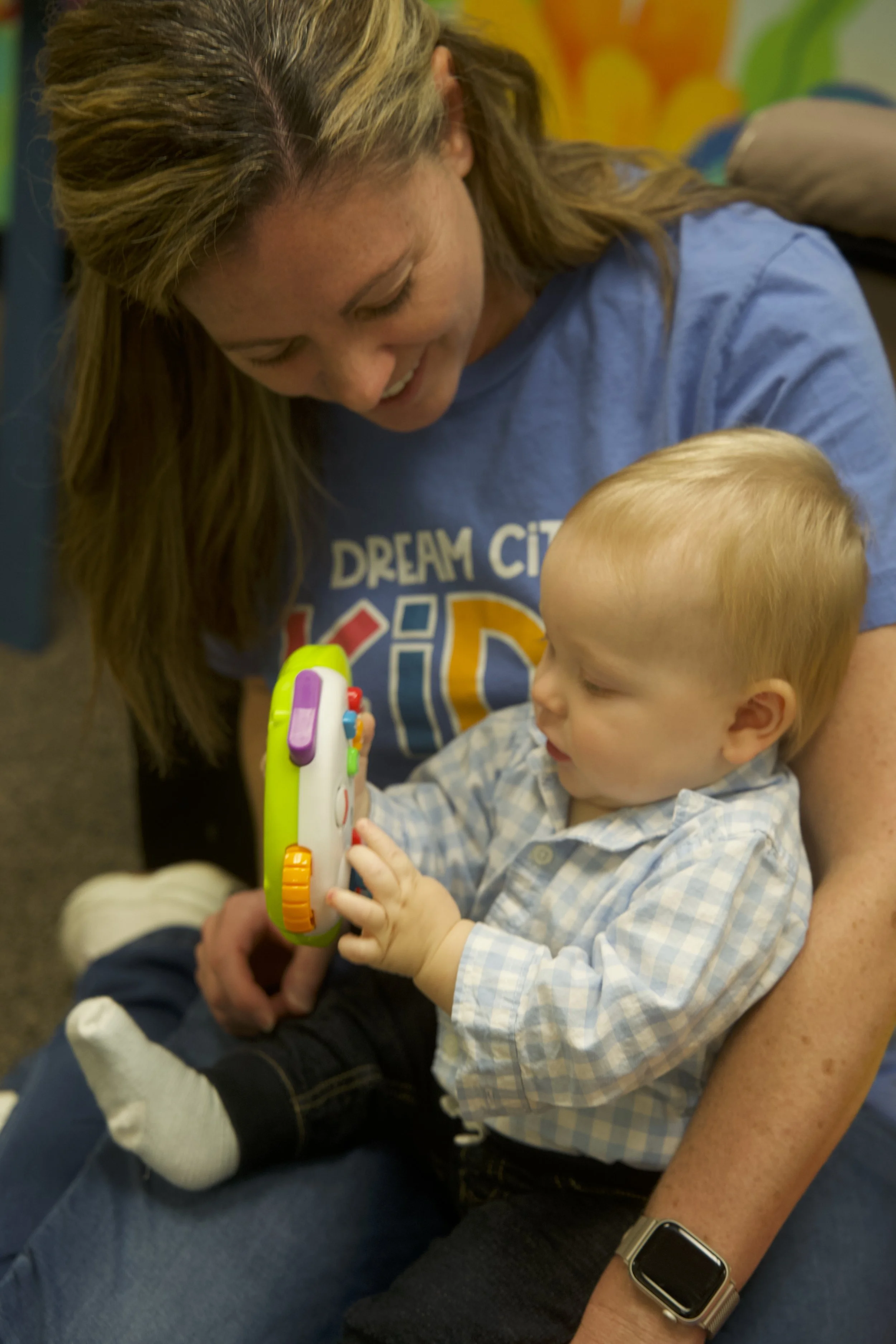 A woman with long blonde hair and a blue t-shirt showing a boy with blonde hair a colorful toy in a preschool or daycare setting.