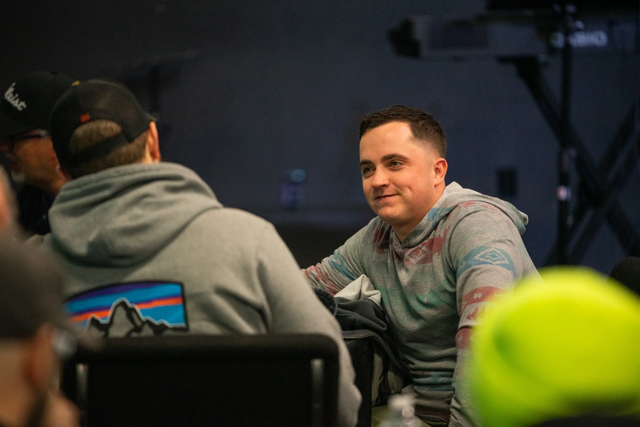 A young man with short dark hair smiling, wearing a gray hoodie with a colorful pattern, sitting at a table engaging in conversation with others, in an indoor setting.