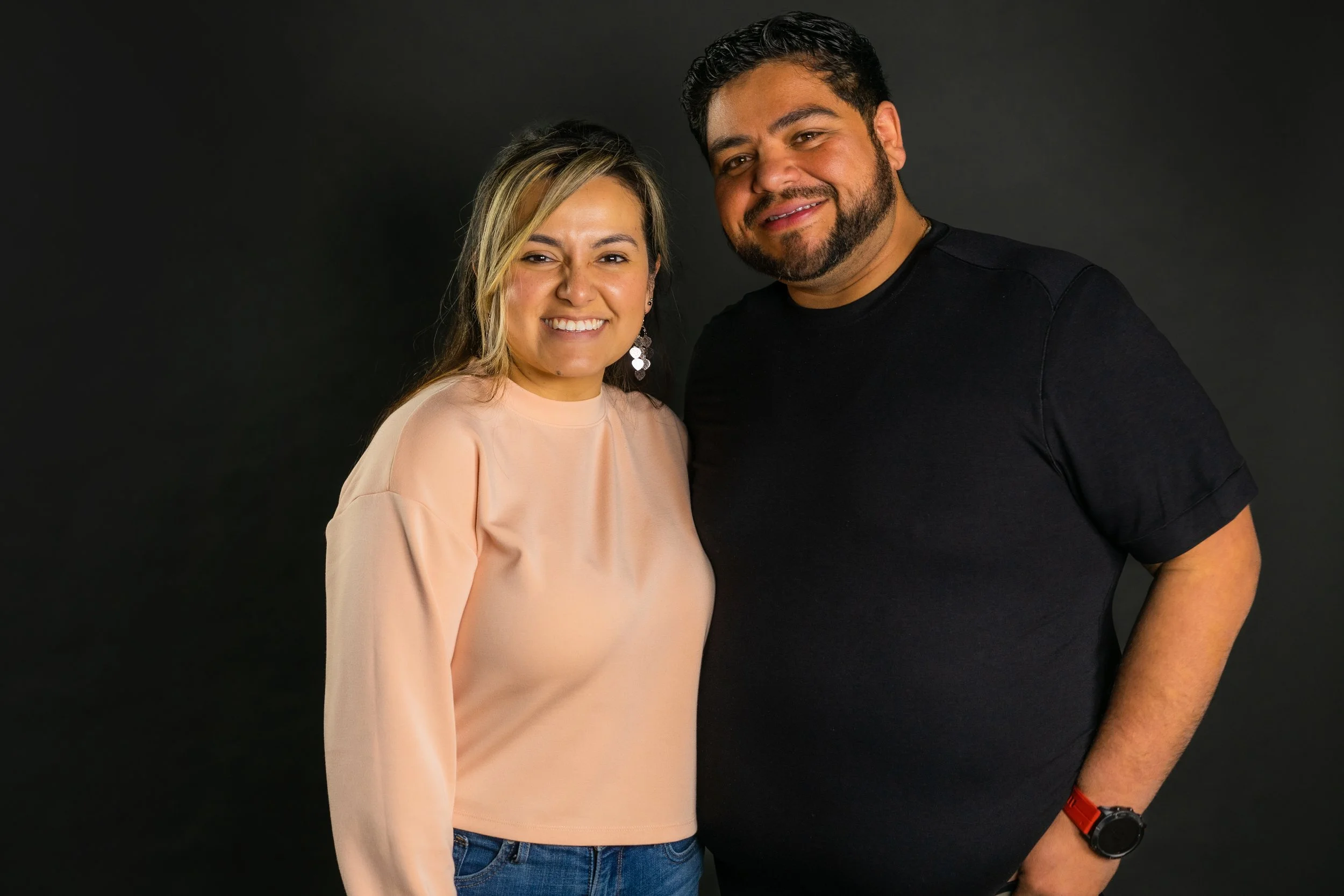 A smiling woman with long hair and earrings standing next to a smiling man with a beard, black t-shirt, and red watch against a dark background.