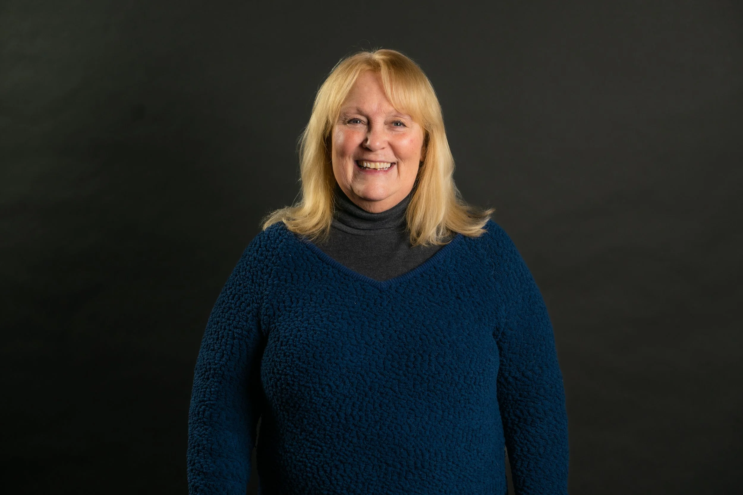 A smiling middle-aged woman with blonde hair, wearing a dark gray turtleneck and a textured blue sweater, standing against a black background.
