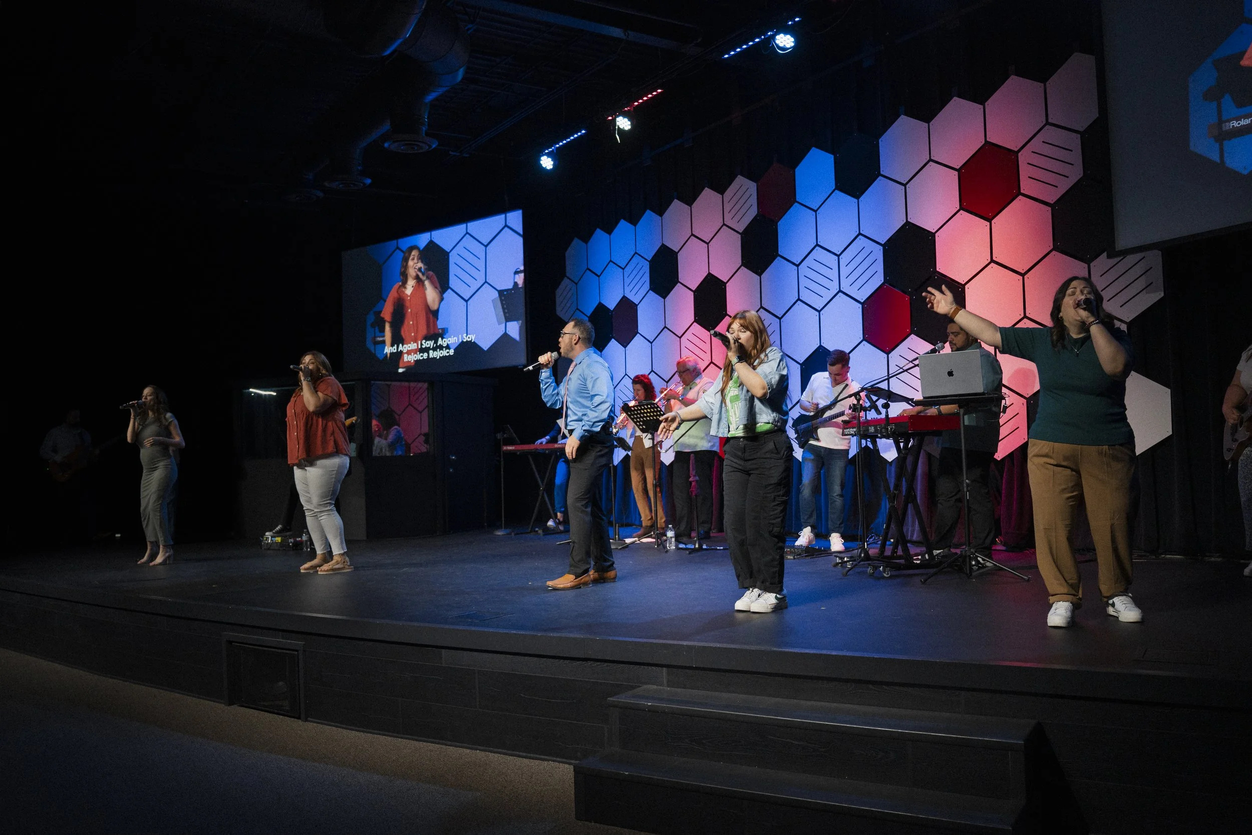 A group of people performing on stage with microphones, musical instruments, and a colorful geometric hexagon backdrop, with a large screen displaying a close-up of a singer.