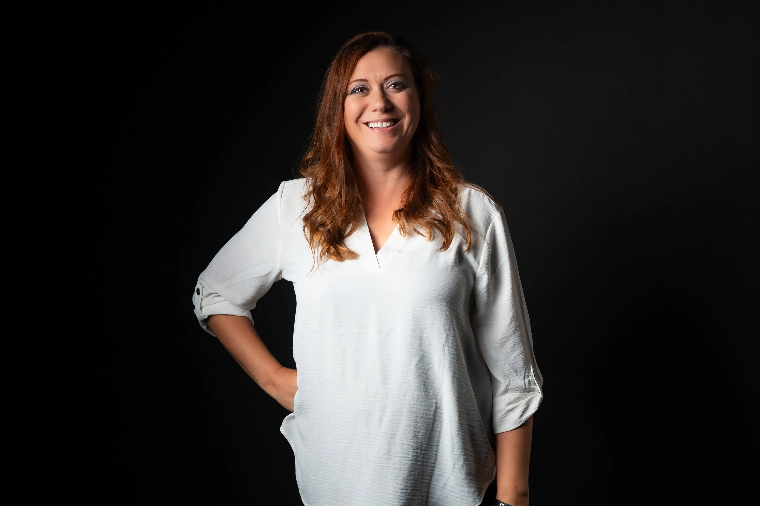 A woman with long, wavy auburn hair, smiling, wearing a white shirt with rolled-up sleeves, standing against a black background.