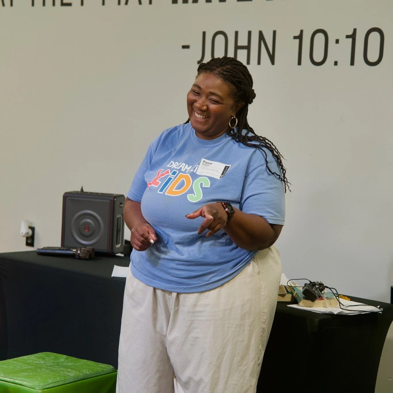 A woman wearing a blue "Dream Kids" T-shirt and beige pants, smiling and pointing with her right hand, standing indoors with a black table behind her, a speaker, papers, and electronic cables on the table, with scripture text on the wall in the background.