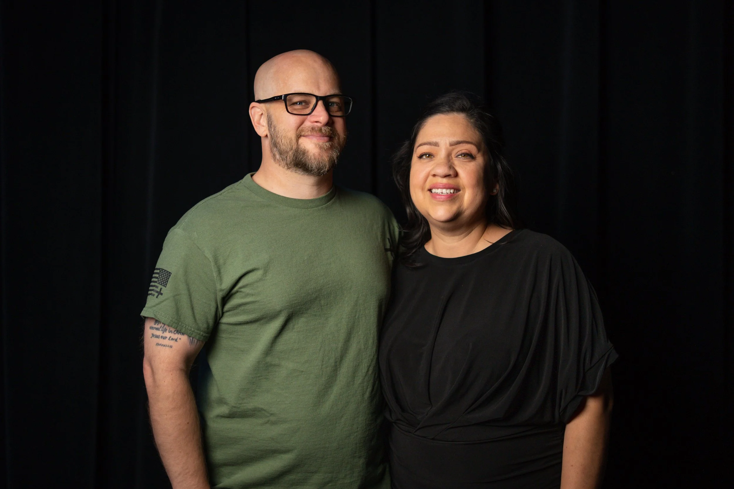 A man with glasses and a beard wearing a green t-shirt, standing next to a woman with black hair wearing a black top, both smiling in front of a black background.