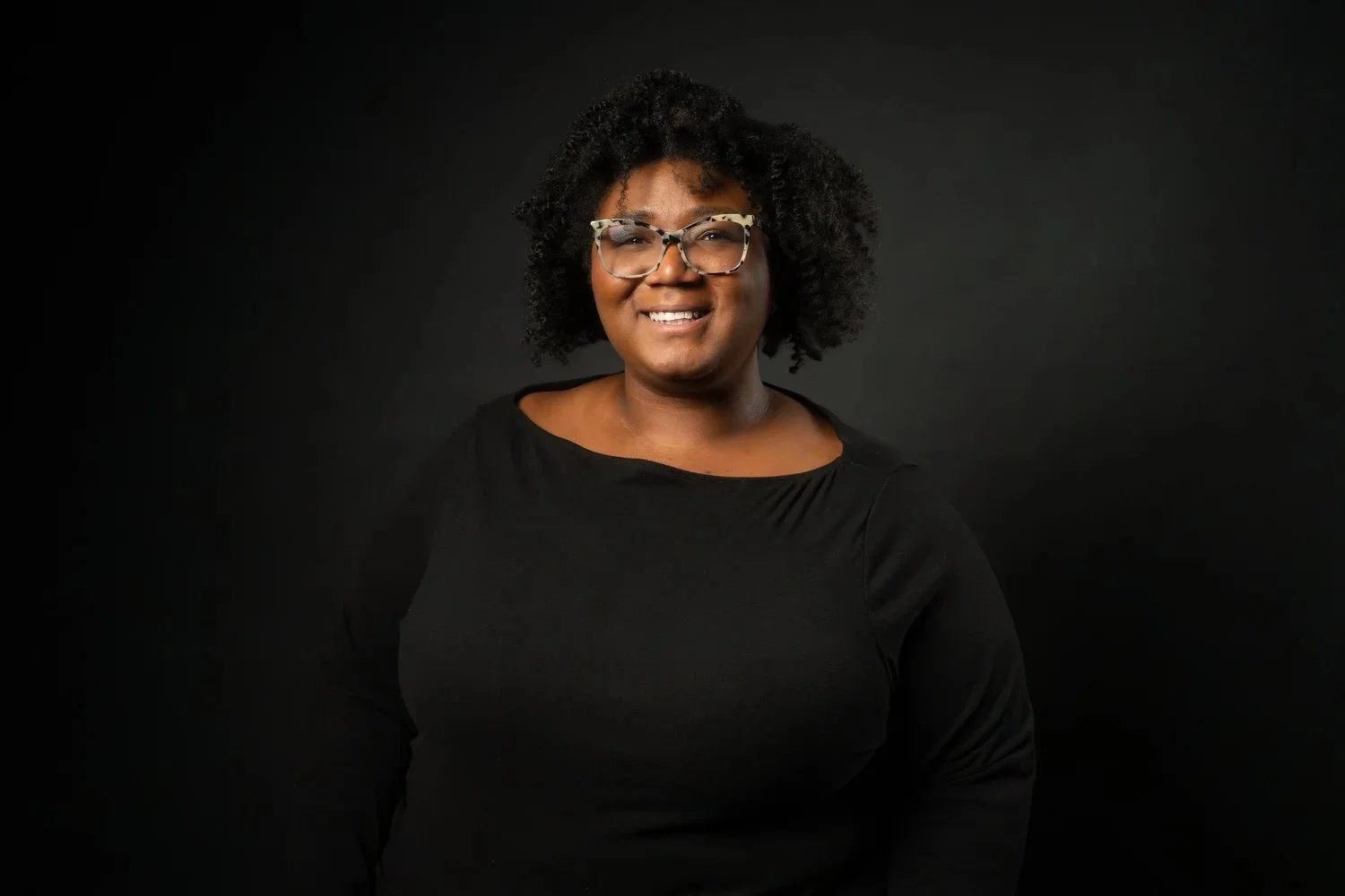A smiling woman with curly hair, glasses, and wearing a black top, against a dark background.
