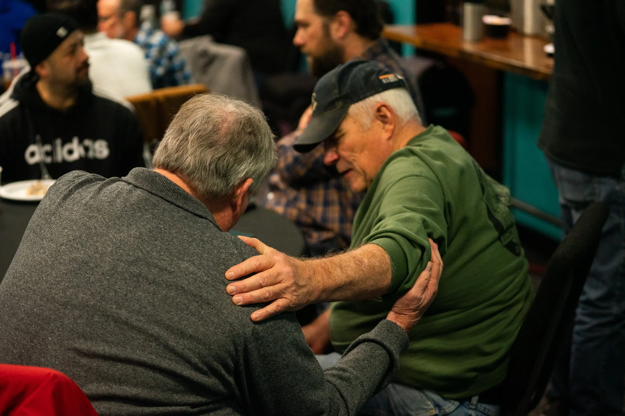 Two elderly men are engaged in a close, comforting conversation. One man is wearing a gray blazer and has his hand on the other's shoulder, expressing concern or support. The other man is wearing a green jacket and a black cap. There are other people in the background, seemingly in a social or casual gathering.