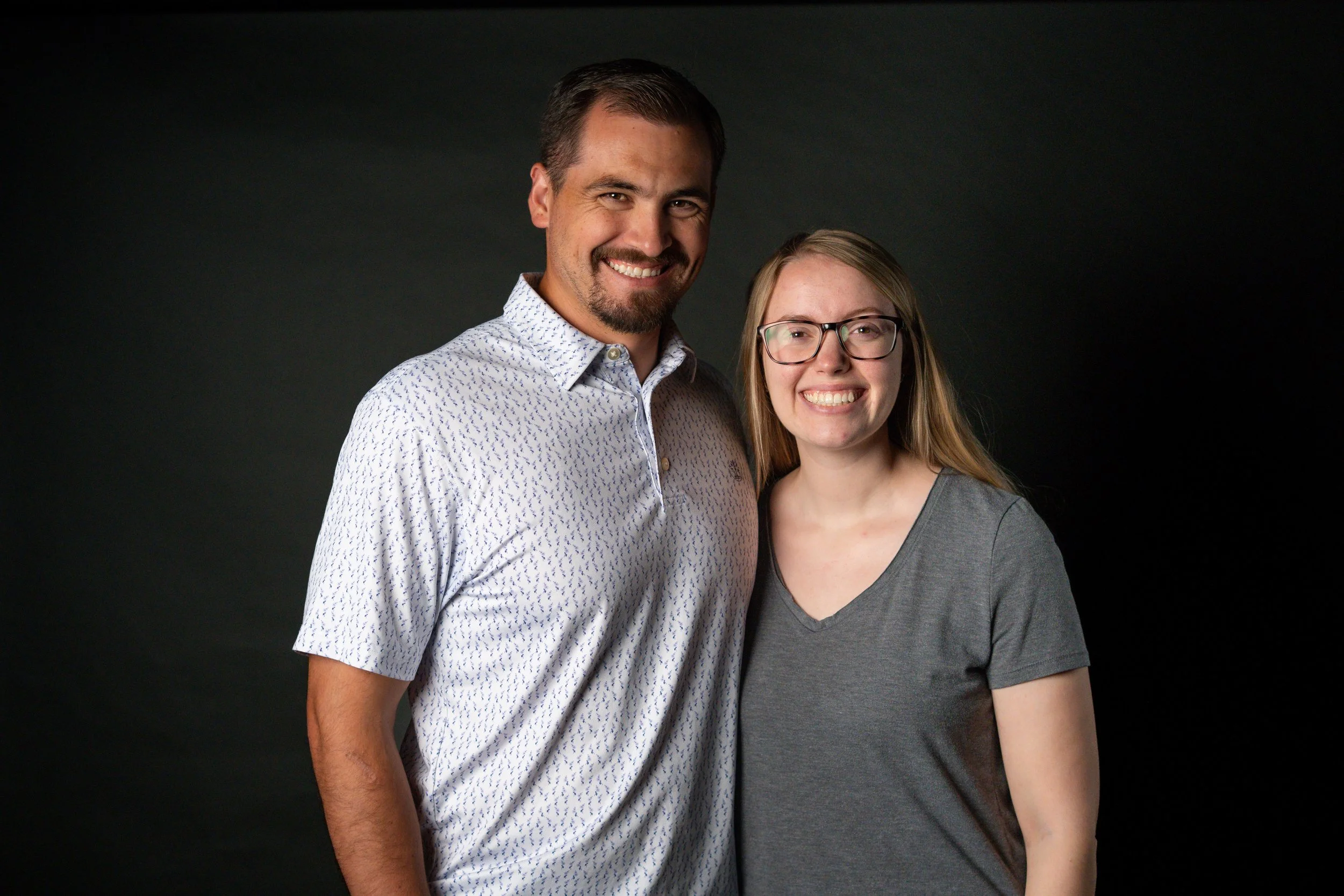 A smiling man and woman standing close together against a dark background, the man wearing a patterned button-up shirt and the woman wearing glasses and a gray T-shirt.