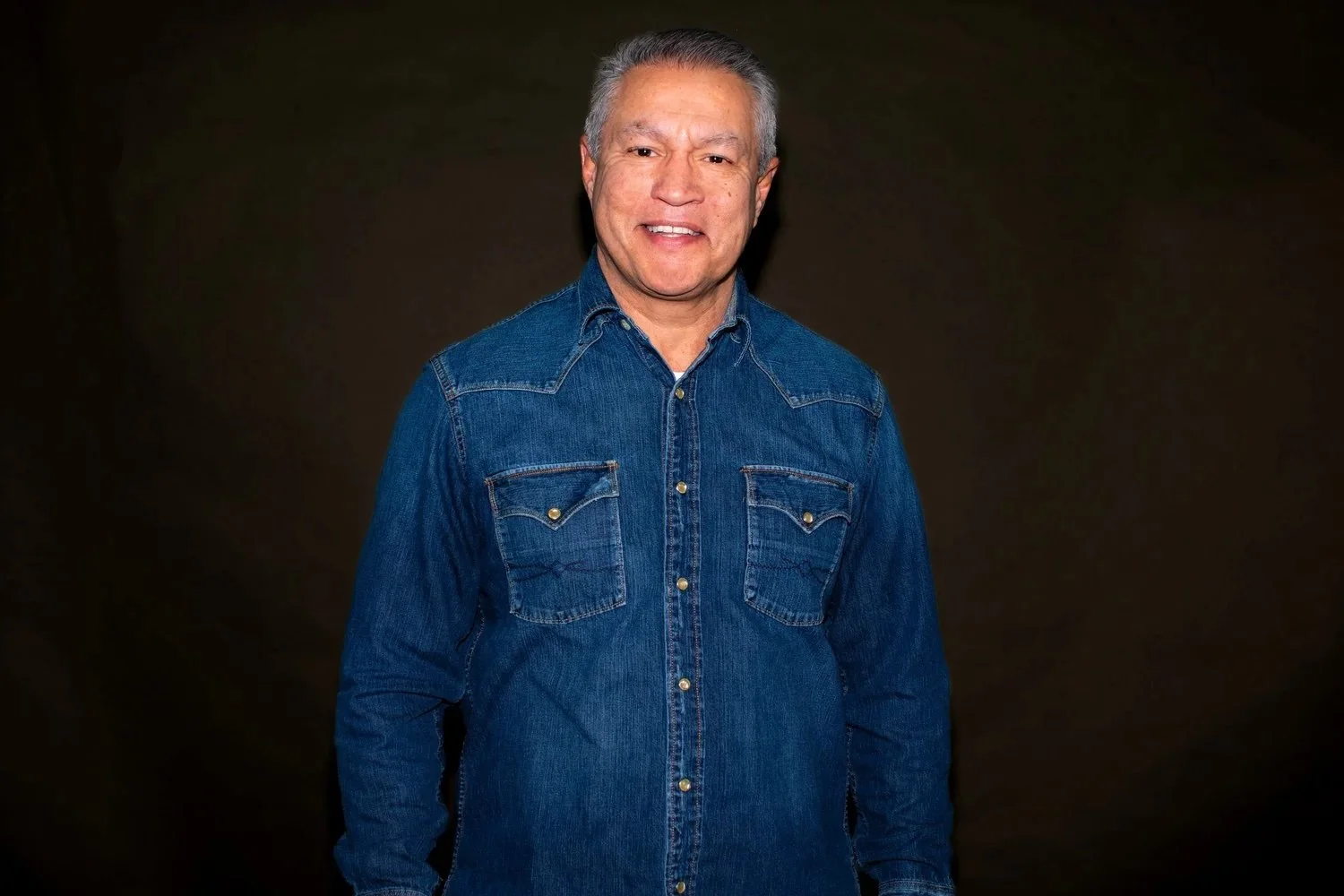 A middle-aged man with gray hair smiling, wearing a blue denim shirt, standing against a black background.