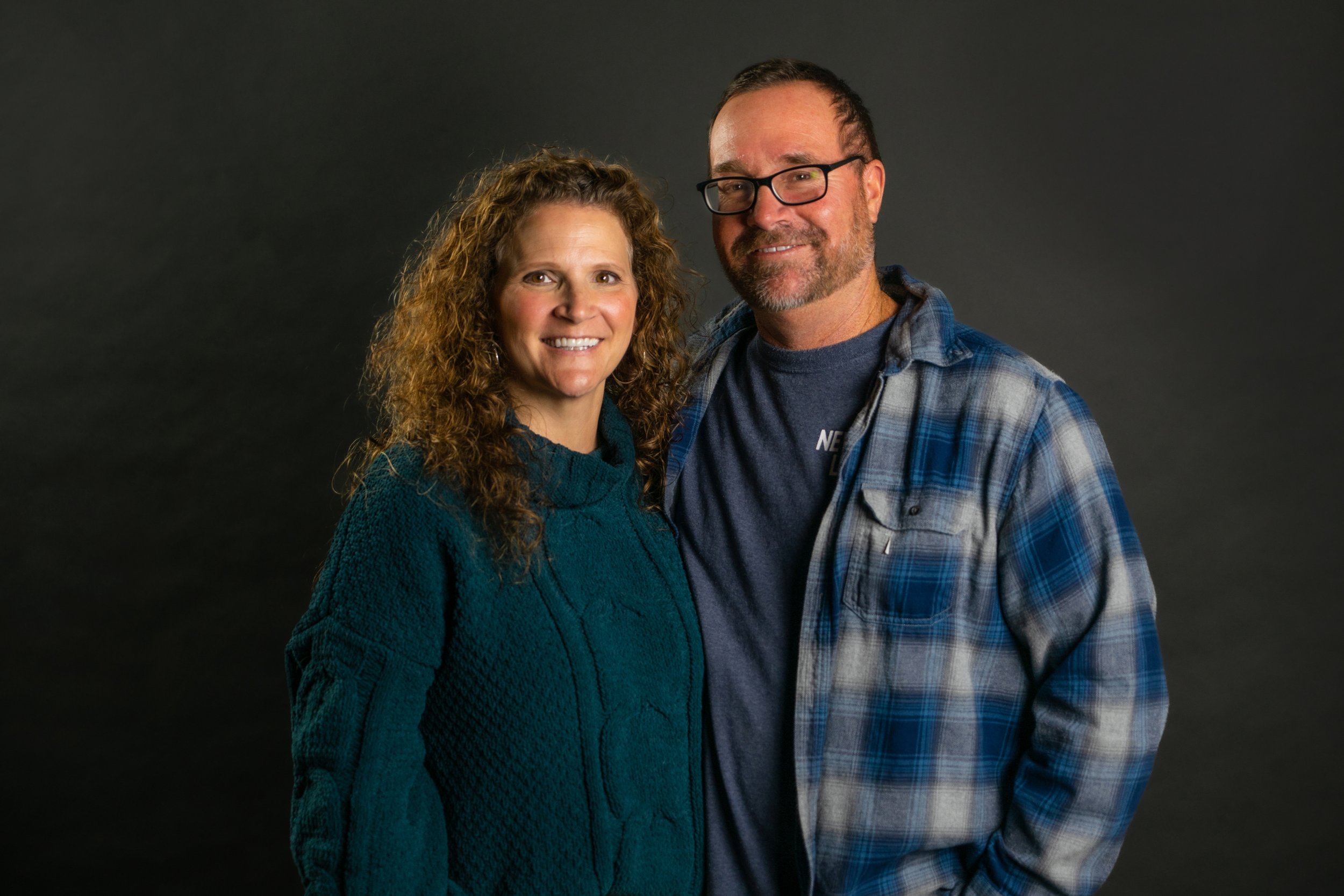 A smiling couple standing close together against a dark background, woman with curly hair wearing a teal sweater, man with glasses and a beard wearing a blue plaid shirt over a T-shirt.