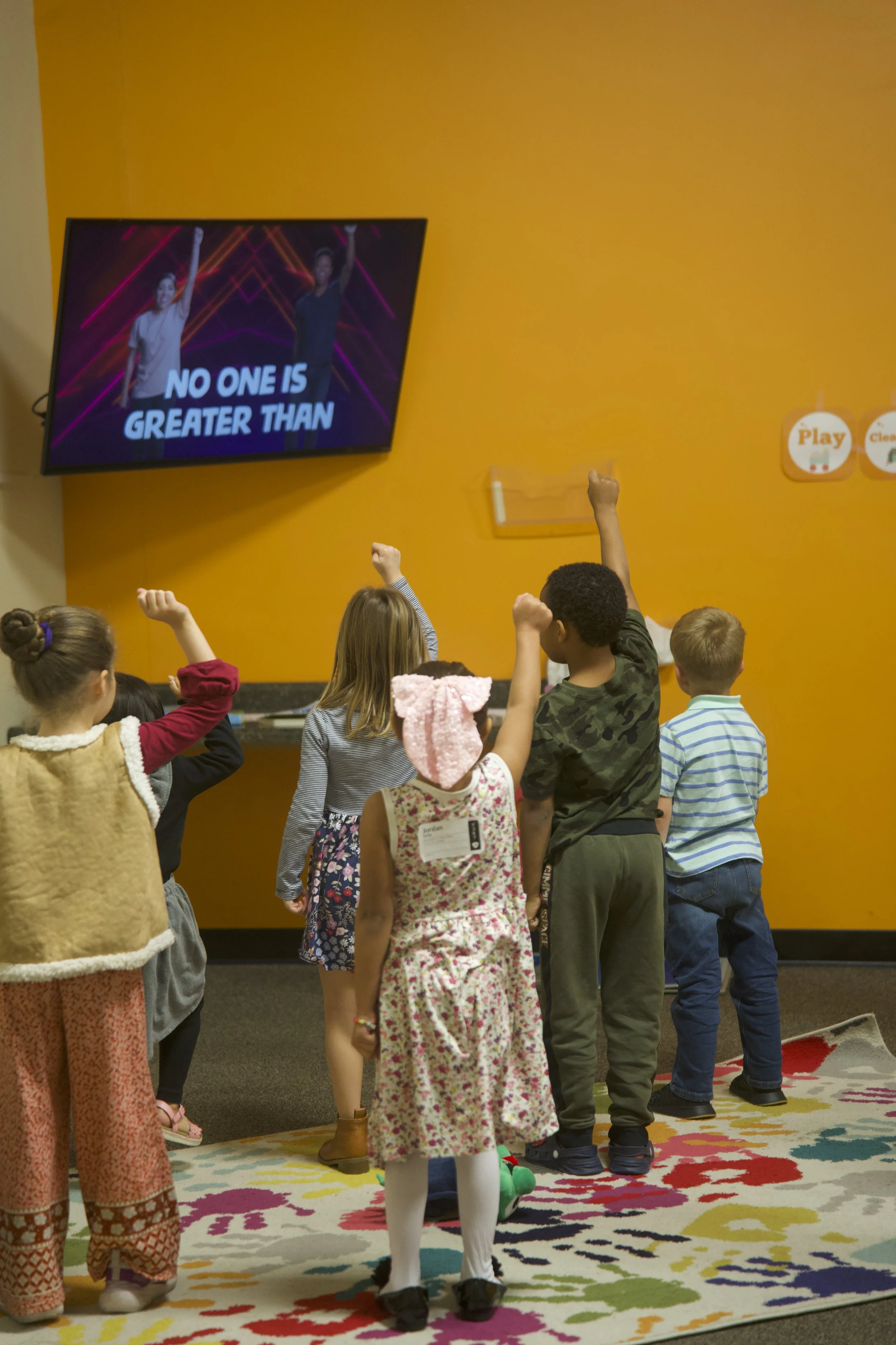Children in a room watching a television screen with a dance or exercise video, raising their fists. The screen displays two people and the text "No one is greater than". The room has an orange wall and a colorful carpet.
