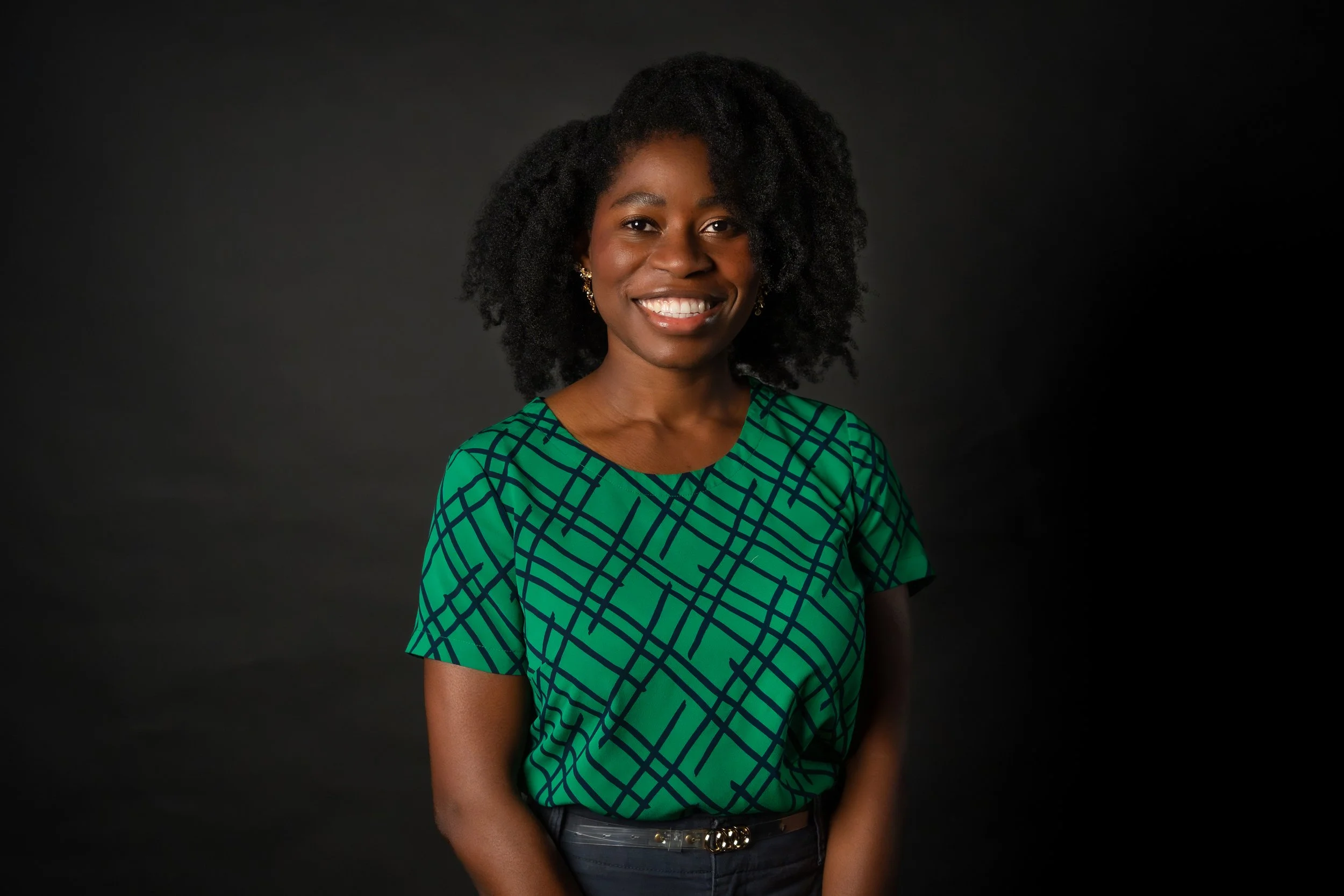 A woman with dark curly hair and a bright smile wearing a green patterned top and earrings against a black background.