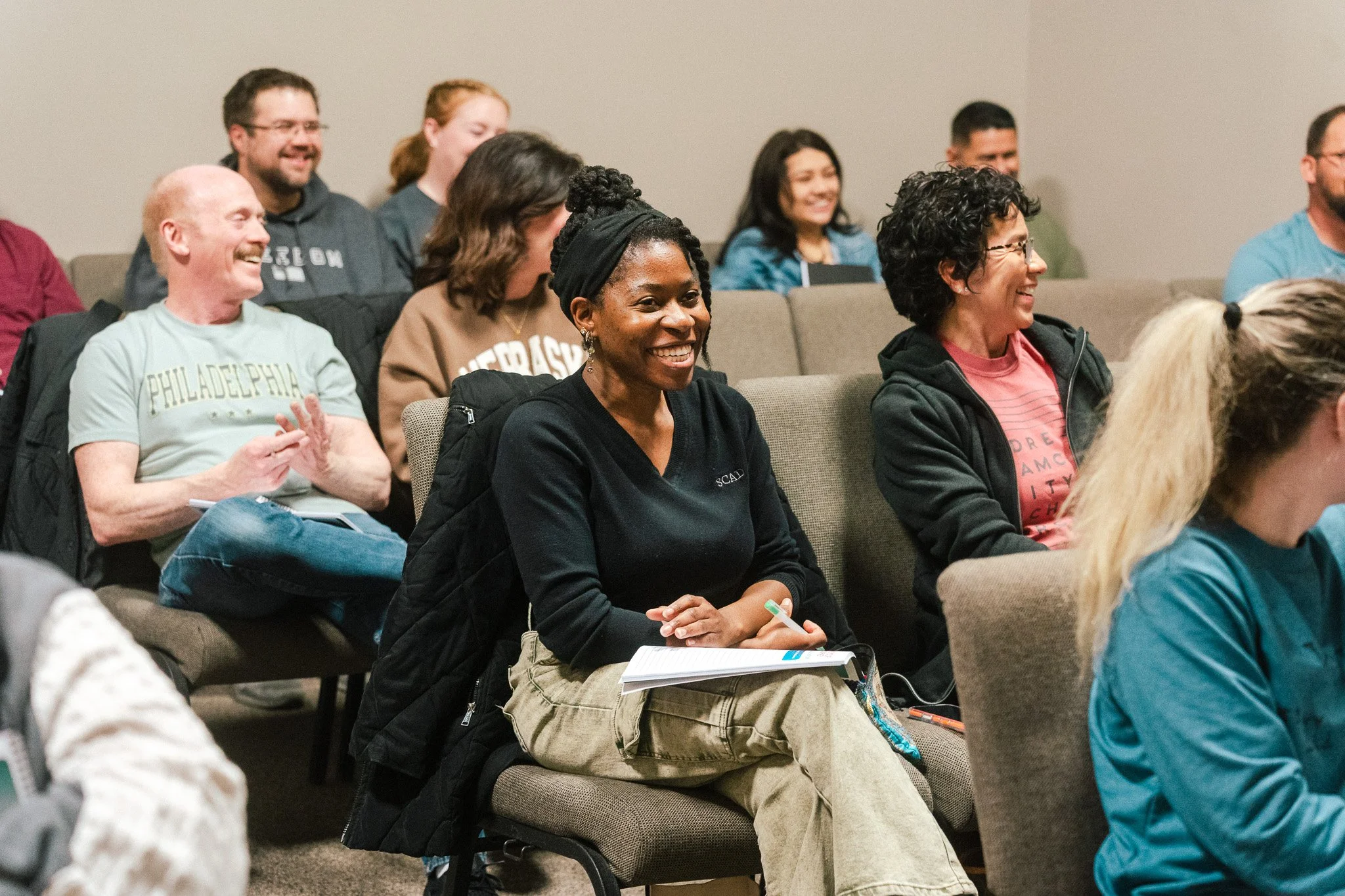 Group of people sitting in chairs at an indoor event, smiling and engaging with a speaker or presentation.