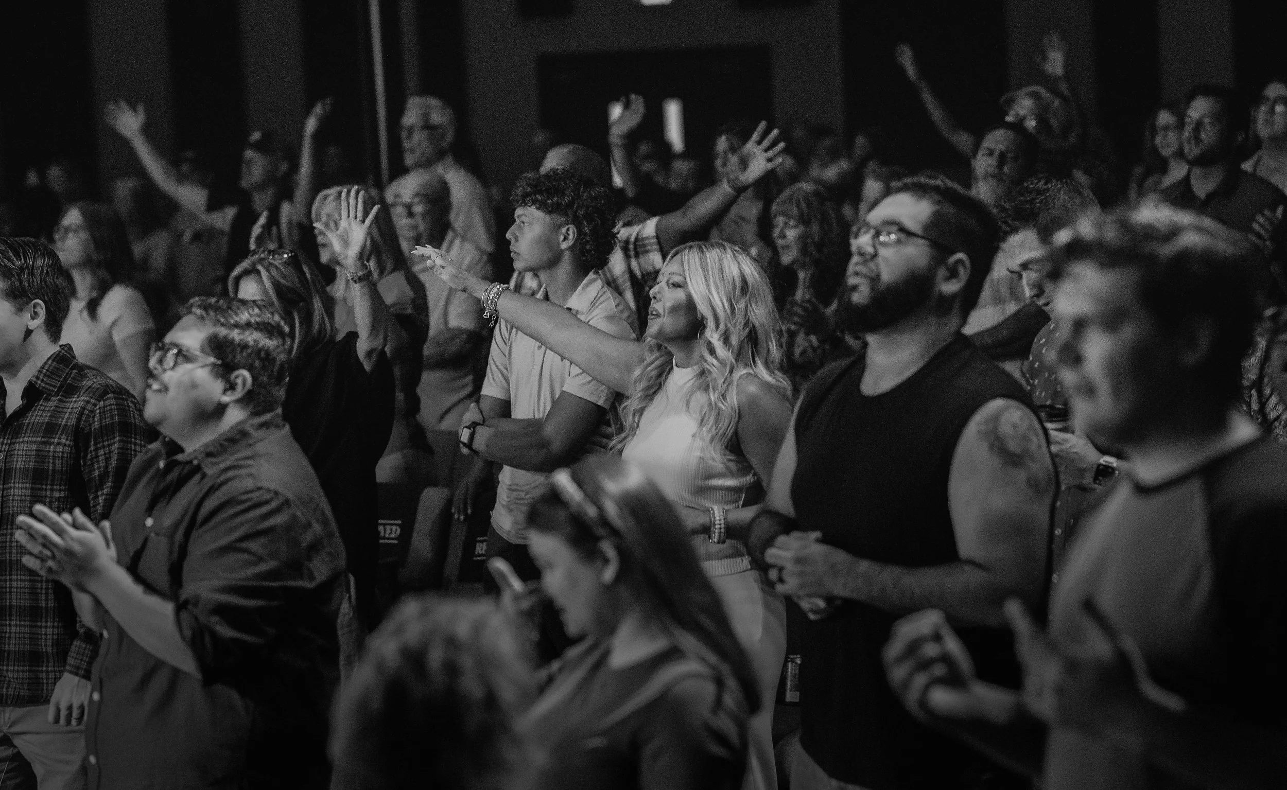 Crowd of diverse people worshiping, some with eyes closed, raising hands, and showing emotional engagement in black and white photo.