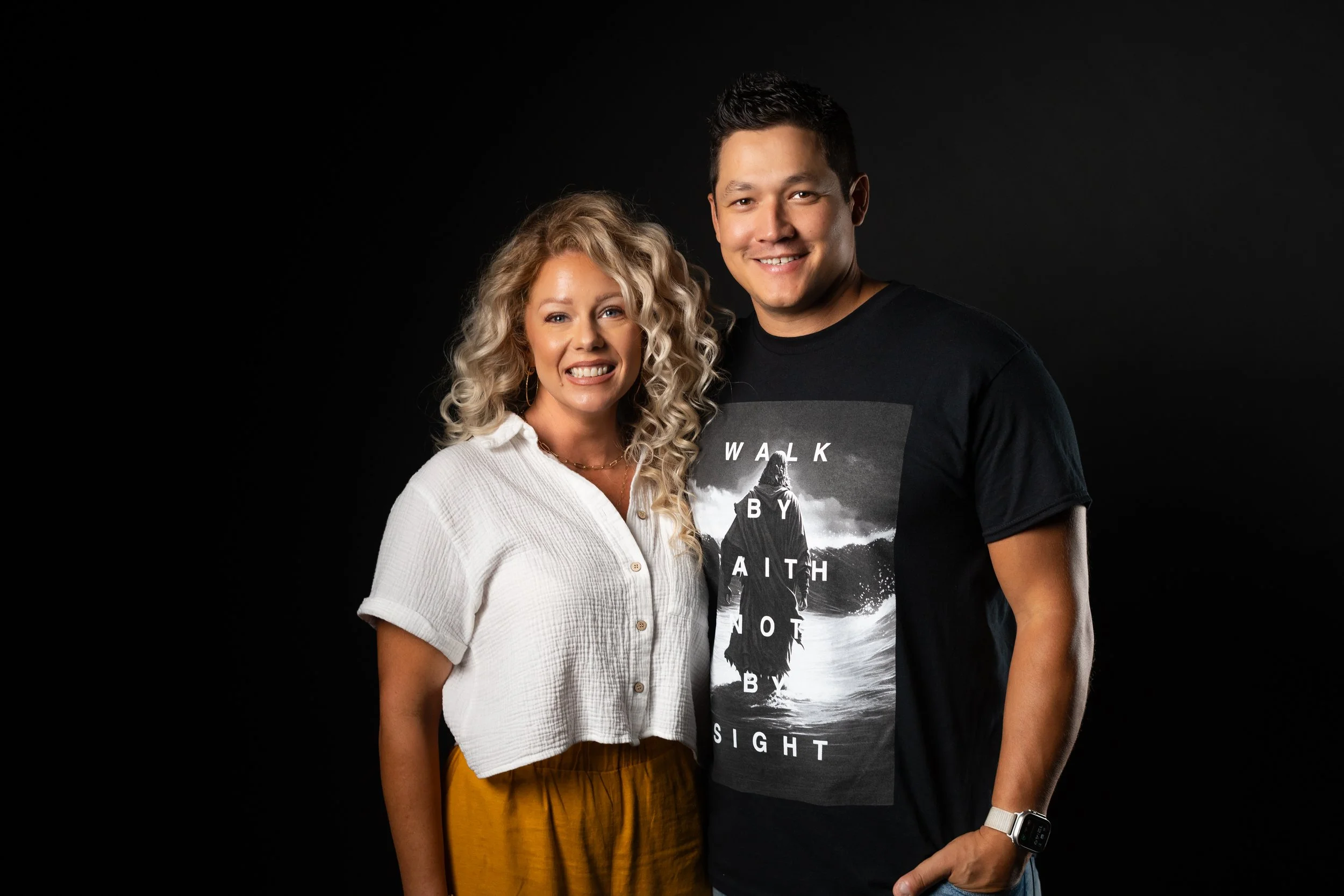 A smiling woman with curly blonde hair wearing a white button-up shirt and mustard-colored skirt standing next to a smiling man with short dark hair wearing a black graphic t-shirt and a white watch, against a black background.