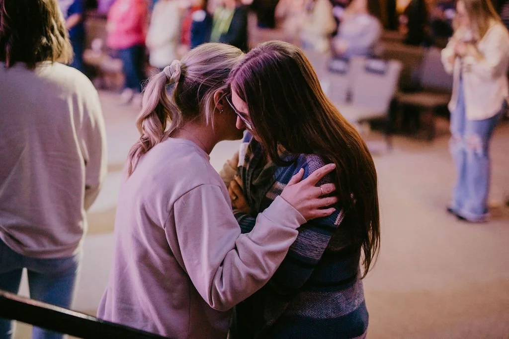 Two women are engaged in a close, emotional moment with their foreheads touching and eyes closed, in a dimly lit, busy indoor setting with other people in the background.