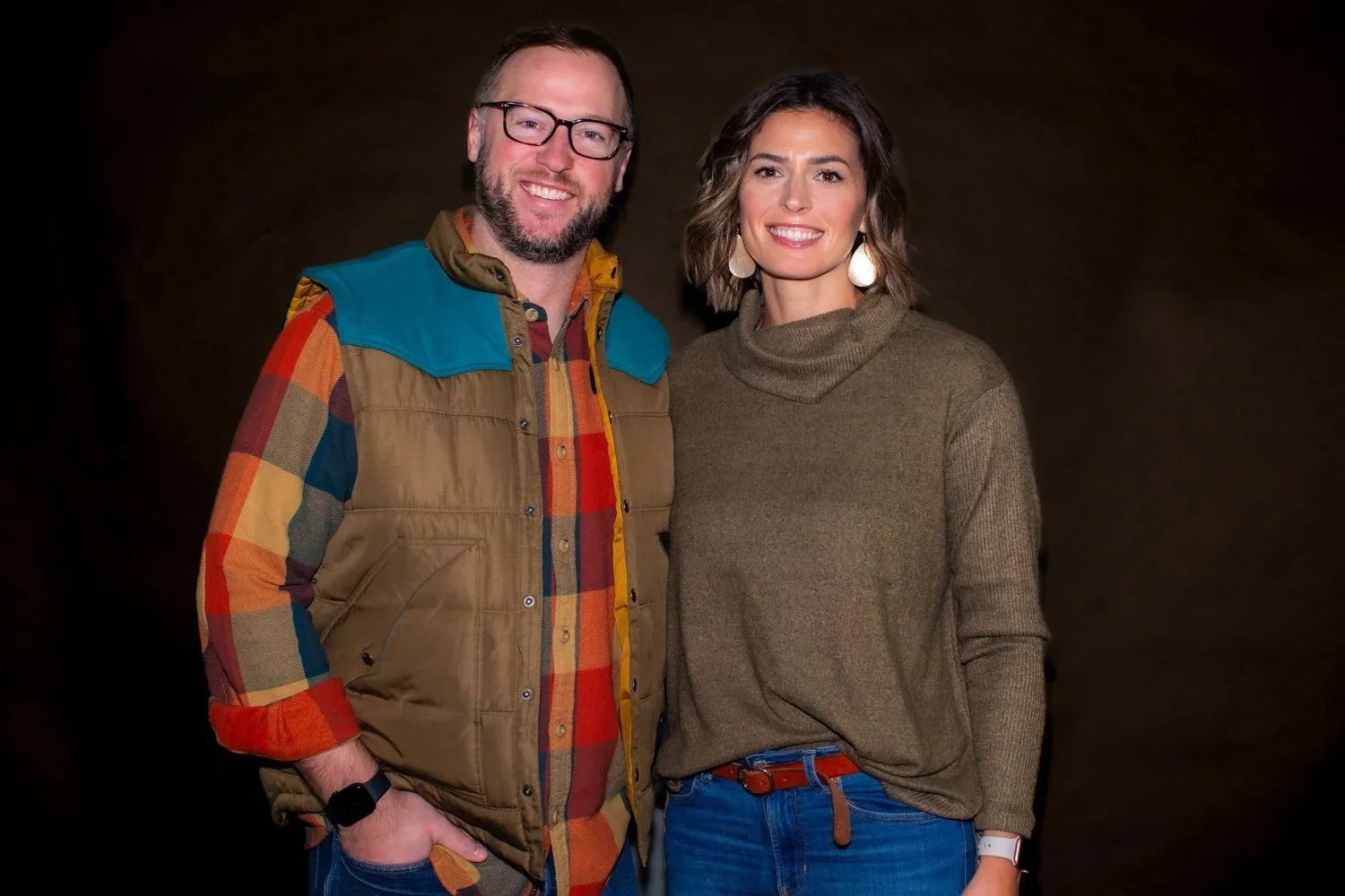 A man and woman standing together, smiling, in front of a dark background. The man wears glasses, a colorful plaid shirt, and a brown vest with a teal shoulder patch. The woman wears a brown turtleneck sweater, blue jeans, and earrings.