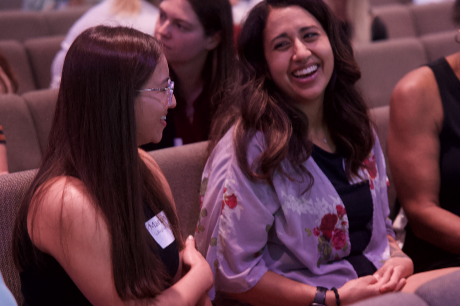 Two women sitting in an audience, smiling and engaging in conversation. One woman has long red hair and glasses, and the other has long brown hair and is wearing a lavender jacket with floral embroidery.