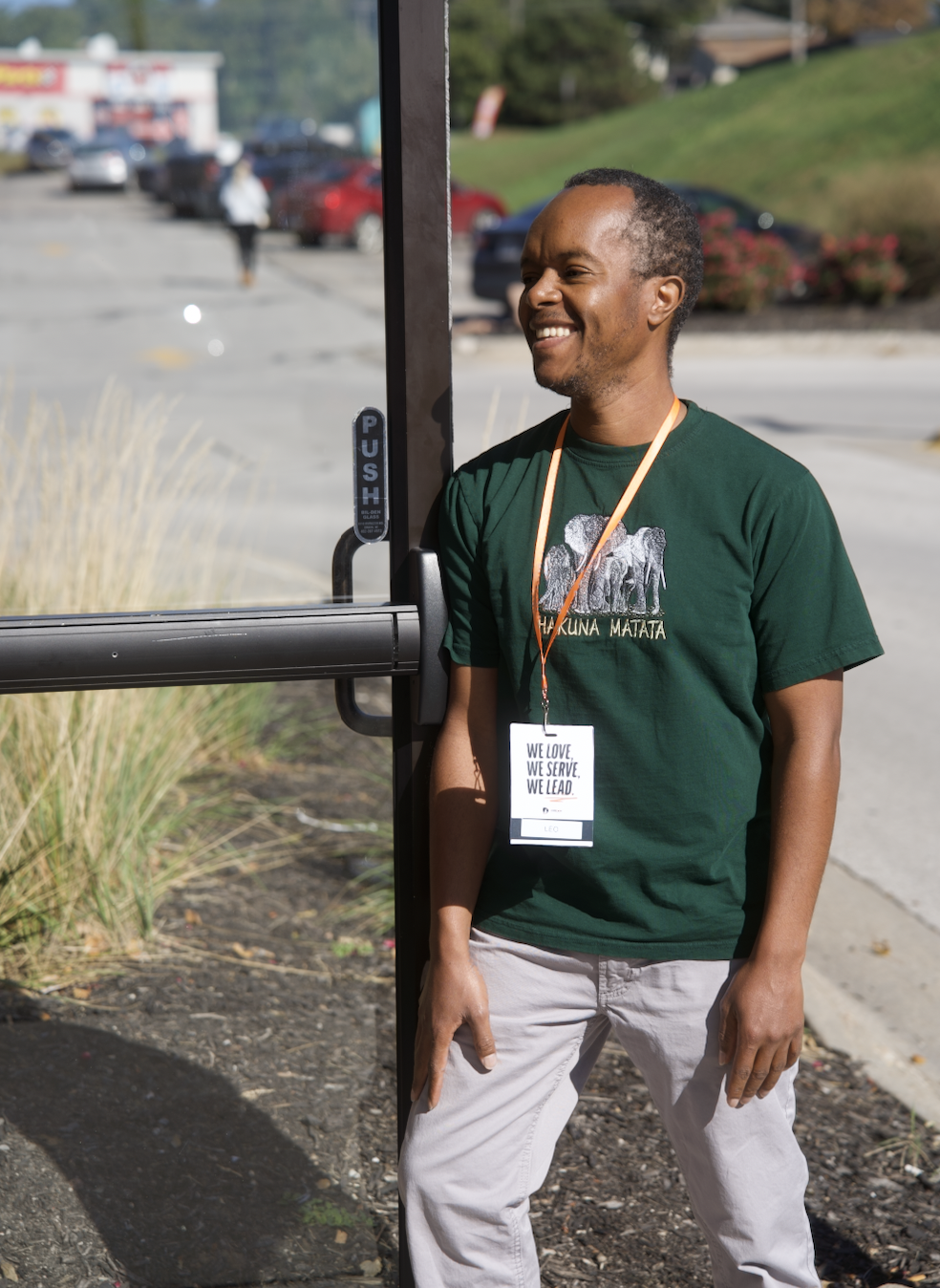 A man standing next to a bus stop shelter on a sunny day, wearing a dark green T-shirt with a lion graphic and the words 'Hakuna Matata', smiling. He's also wearing a name tag that says 'Leo' and a lanyard.