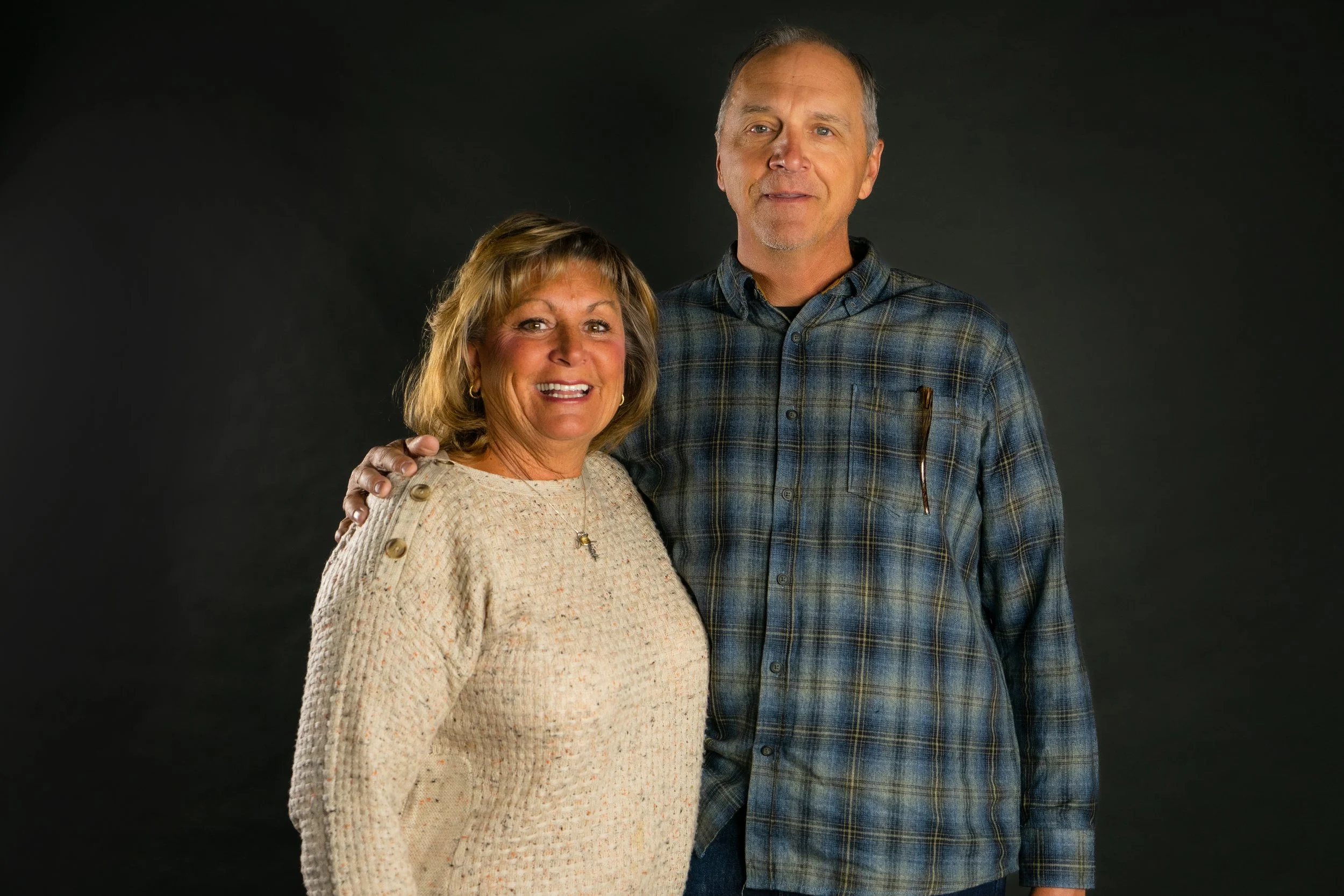 A smiling woman with blonde hair and gold earrings wearing a beige sweater standing next to a taller man with short gray hair, wearing a blue plaid shirt against a black background.
