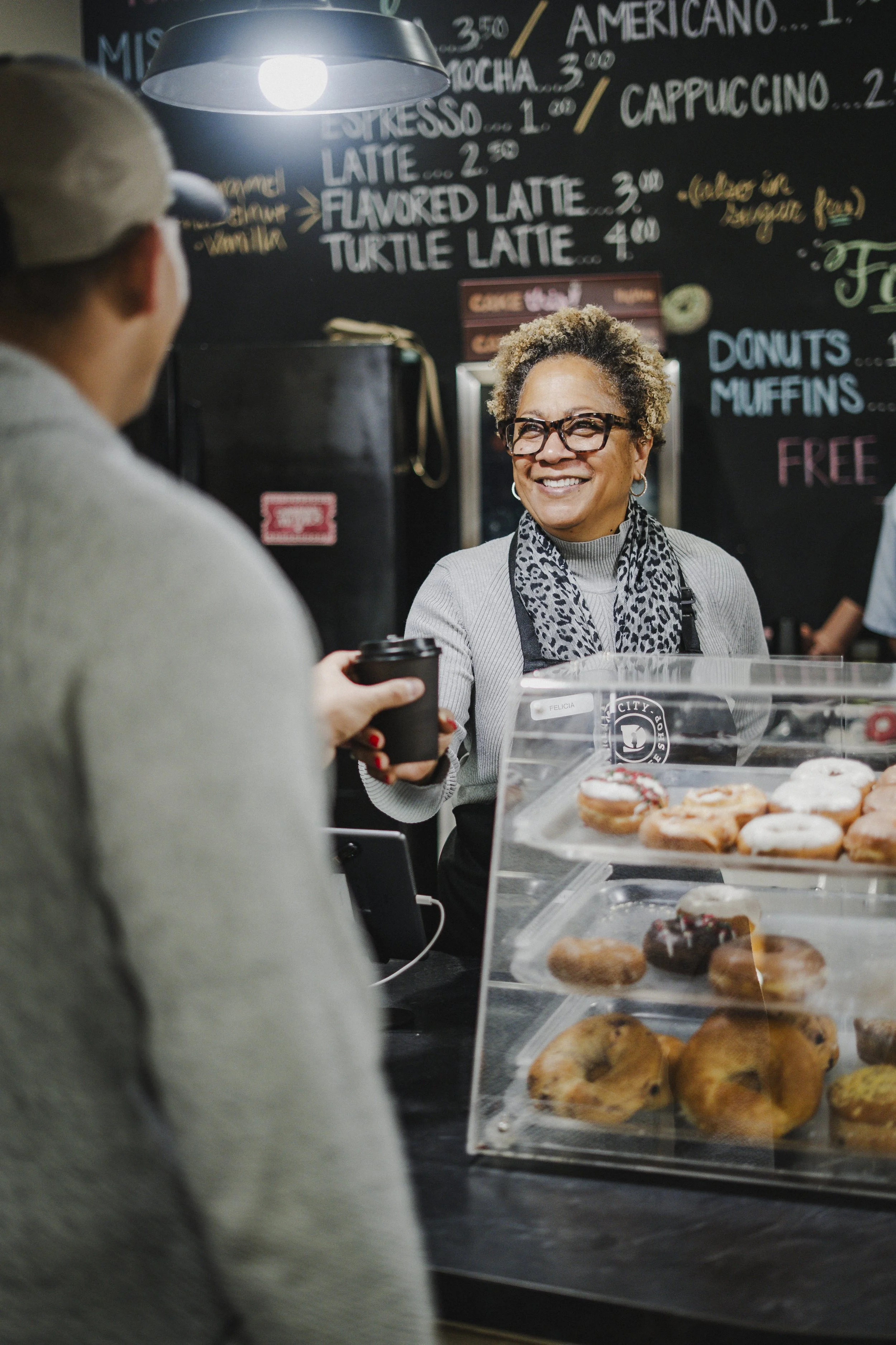 A smiling woman working at a bakery counter receives a coffee from a customer. A display case with various donuts is visible in the foreground, and a menu board with beverage options is in the background.