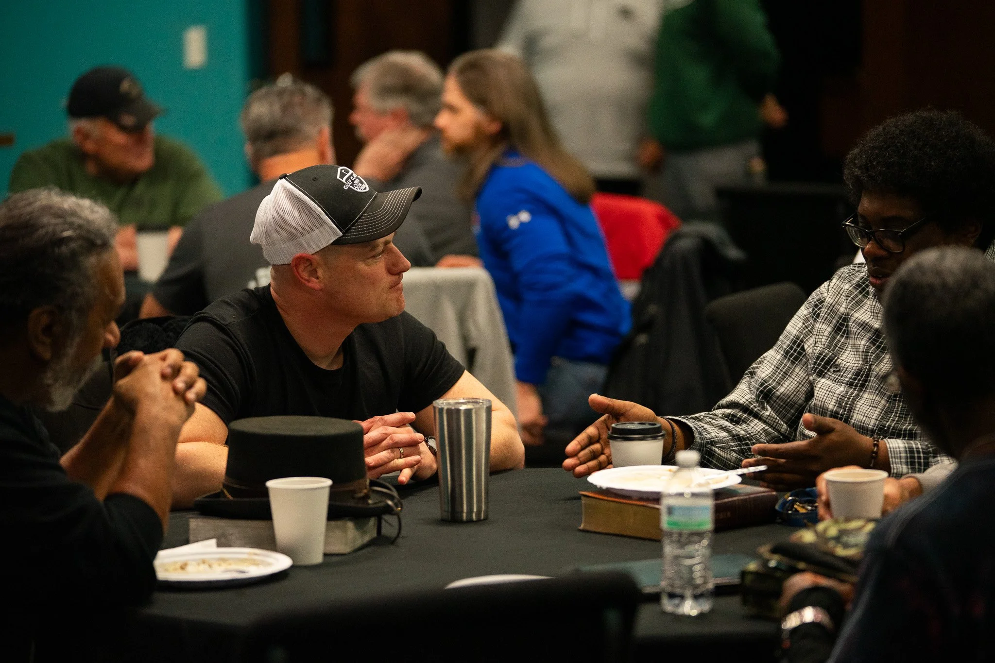 People sitting at a table engaged in conversation, with plates, cups, and bottled water on the table, in a dimly lit indoor setting.