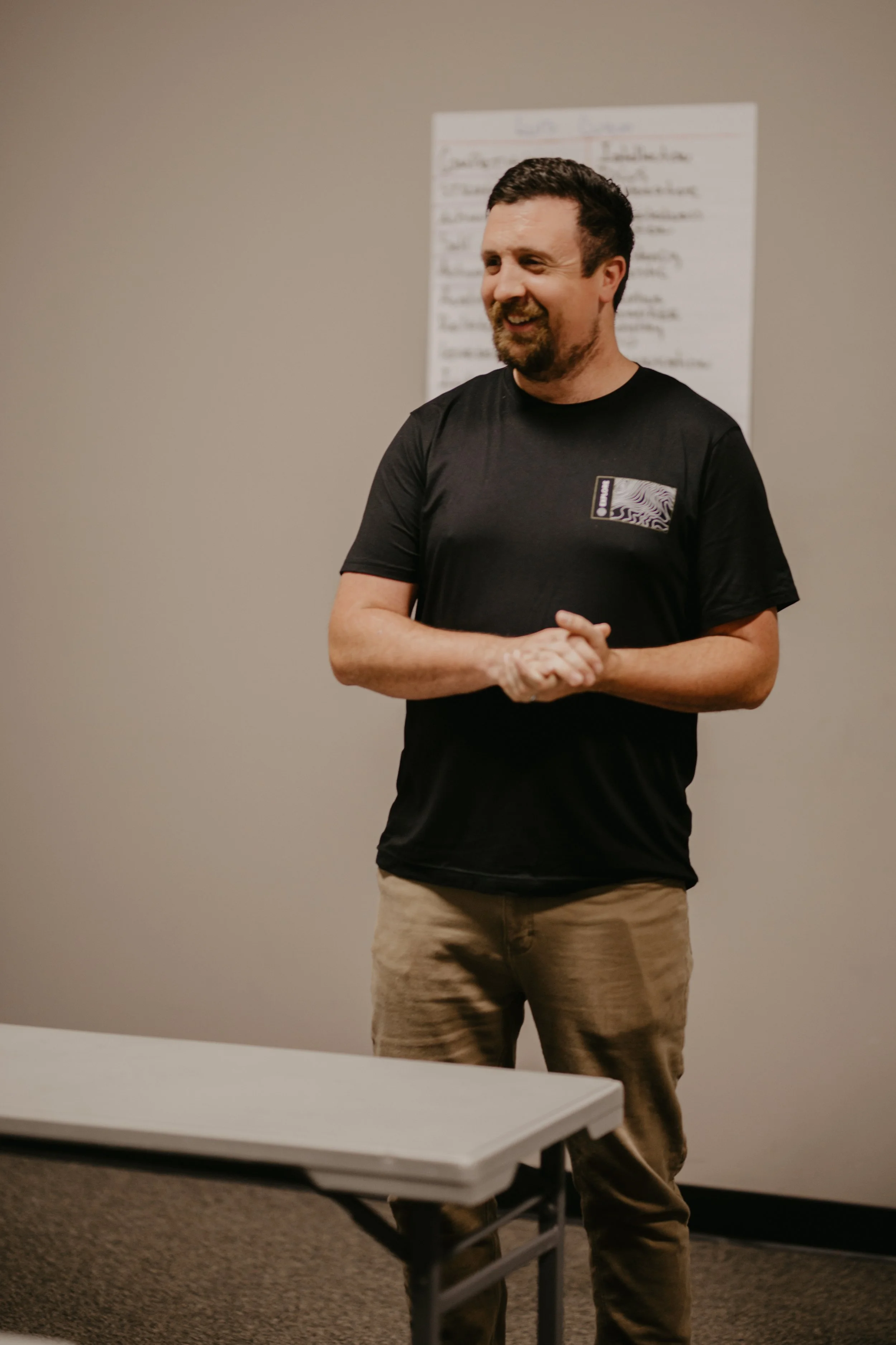 A man is standing and smiling in a room, wearing a black T-shirt and khaki pants, with a whiteboard in the background.
