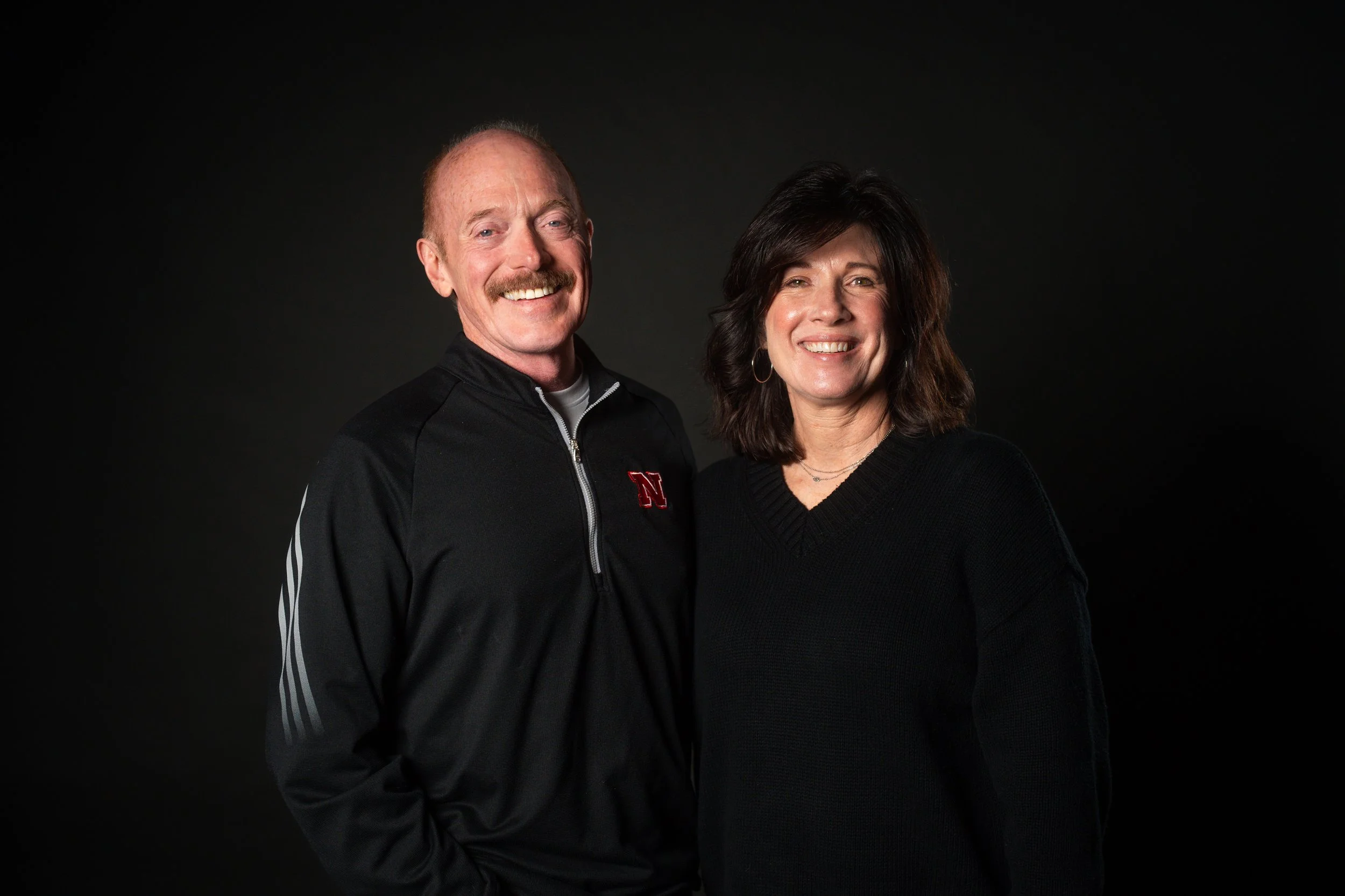 A smiling man with a mustache wearing a black Nebraska sports jacket standing next to a smiling woman with brown hair wearing a black sweater against a dark background.
