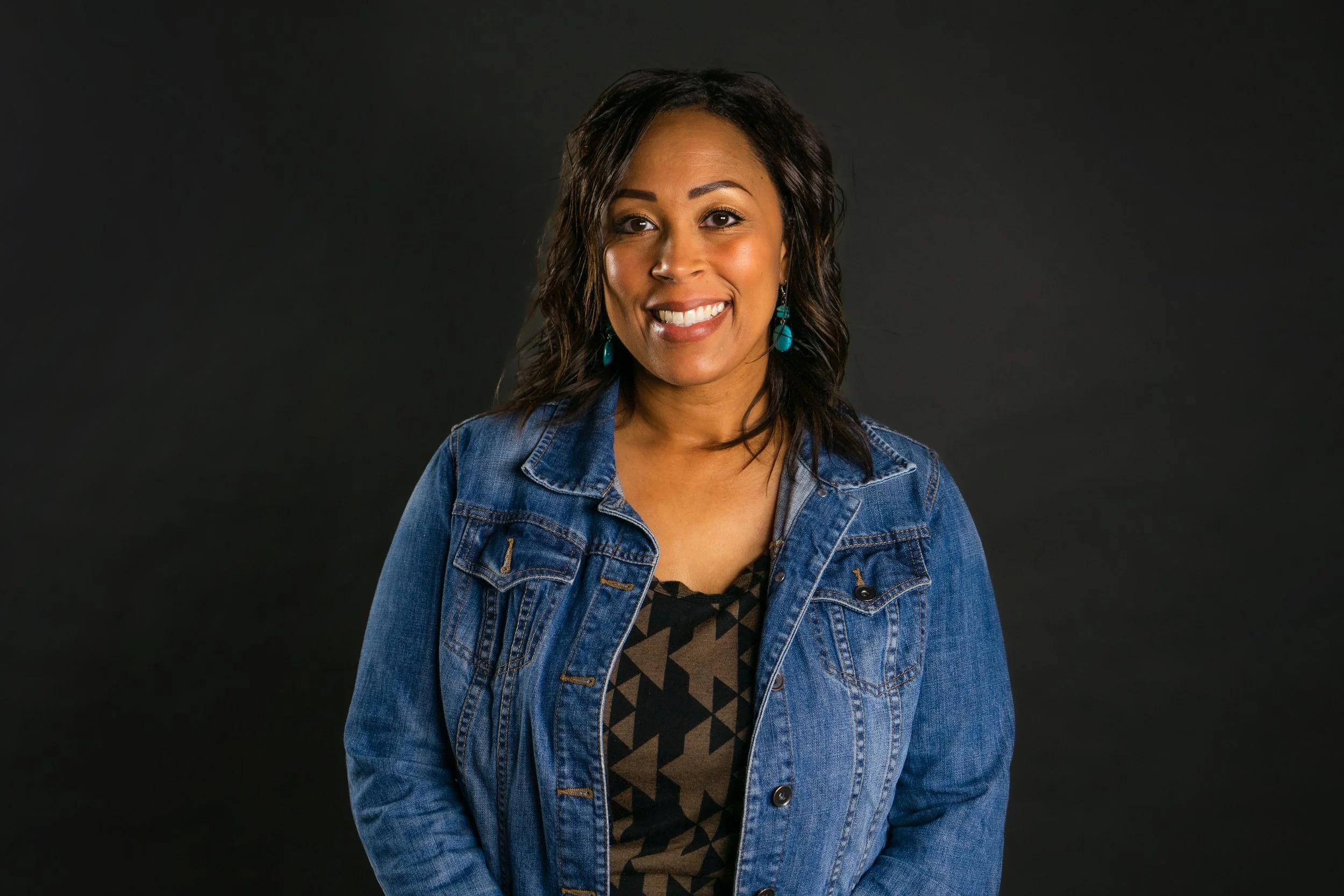 A woman with shoulder-length dark hair, smiling, wearing turquoise earrings, a patterned shirt, and a denim jacket against a black background.