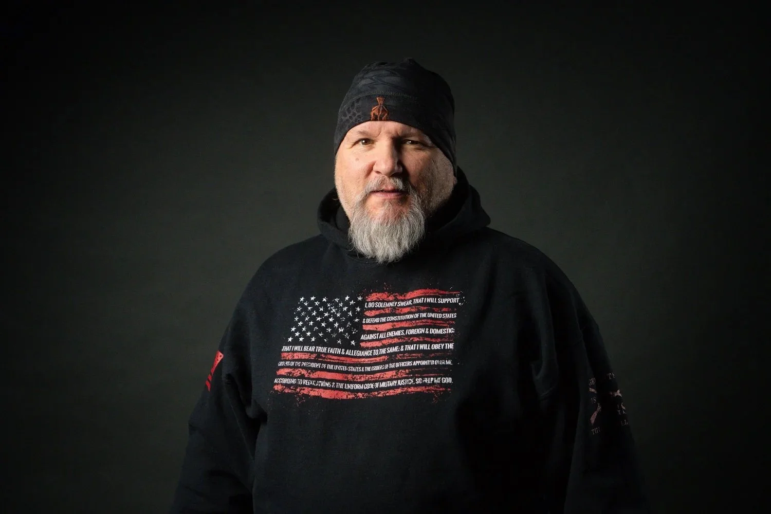 A man with a gray beard wears a black beanie and black hoodie with an American flag and text. He looks at the camera with a neutral expression, against a dark background.