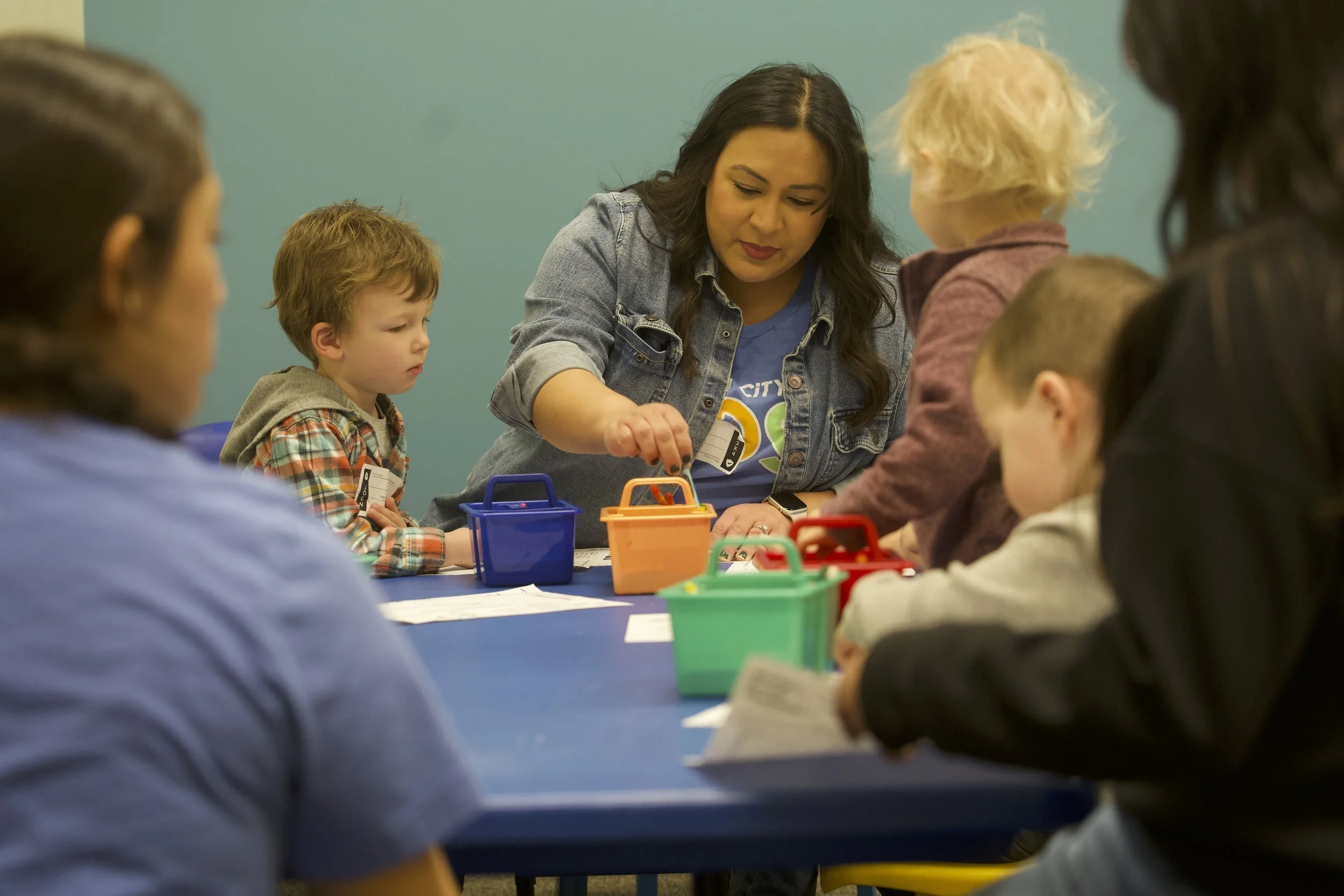 A woman is leading a group activity with children at a table, with colorful containers and papers, in a classroom setting.