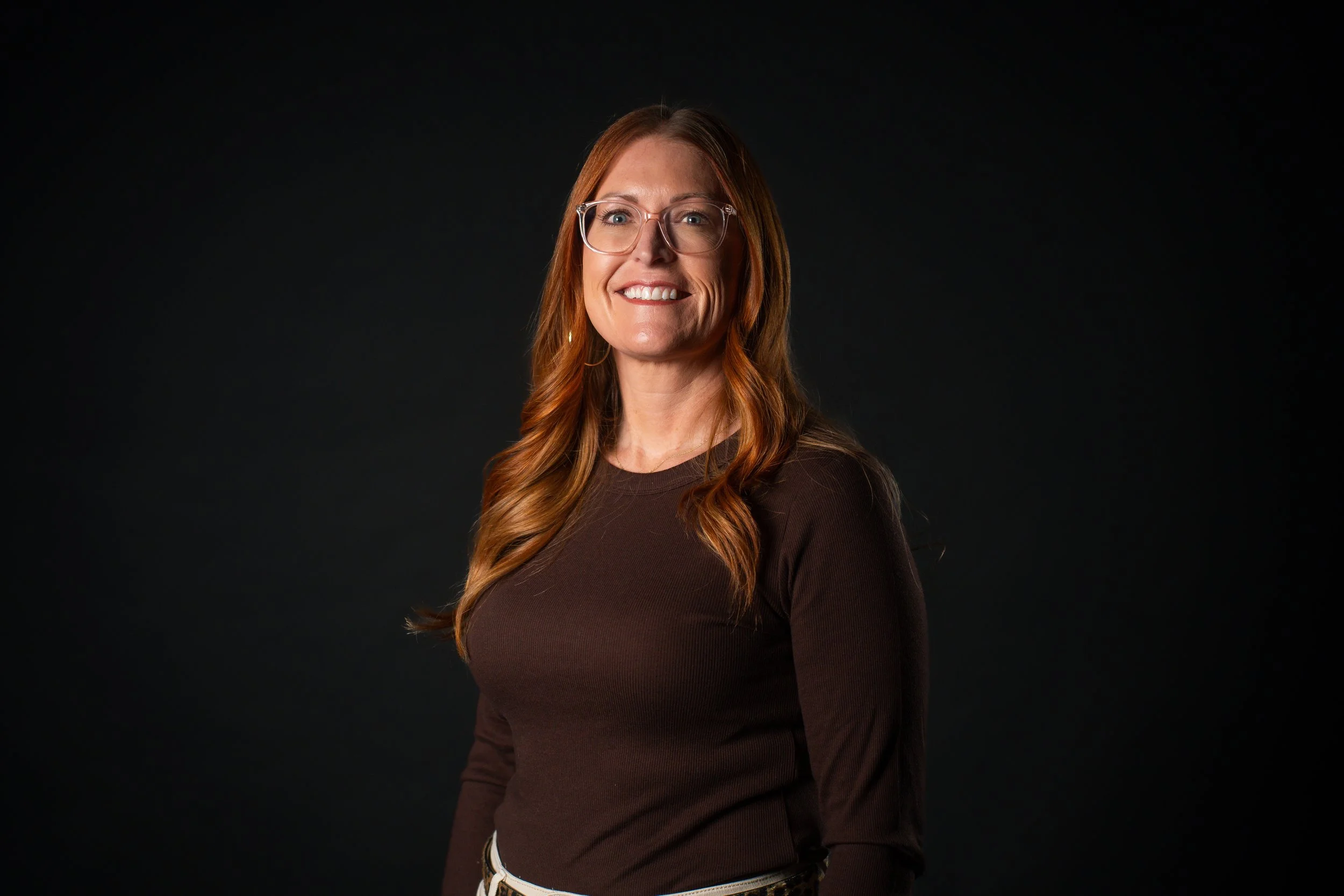A woman with long red hair, glasses, and a brown long-sleeve shirt standing against a black background, smiling.