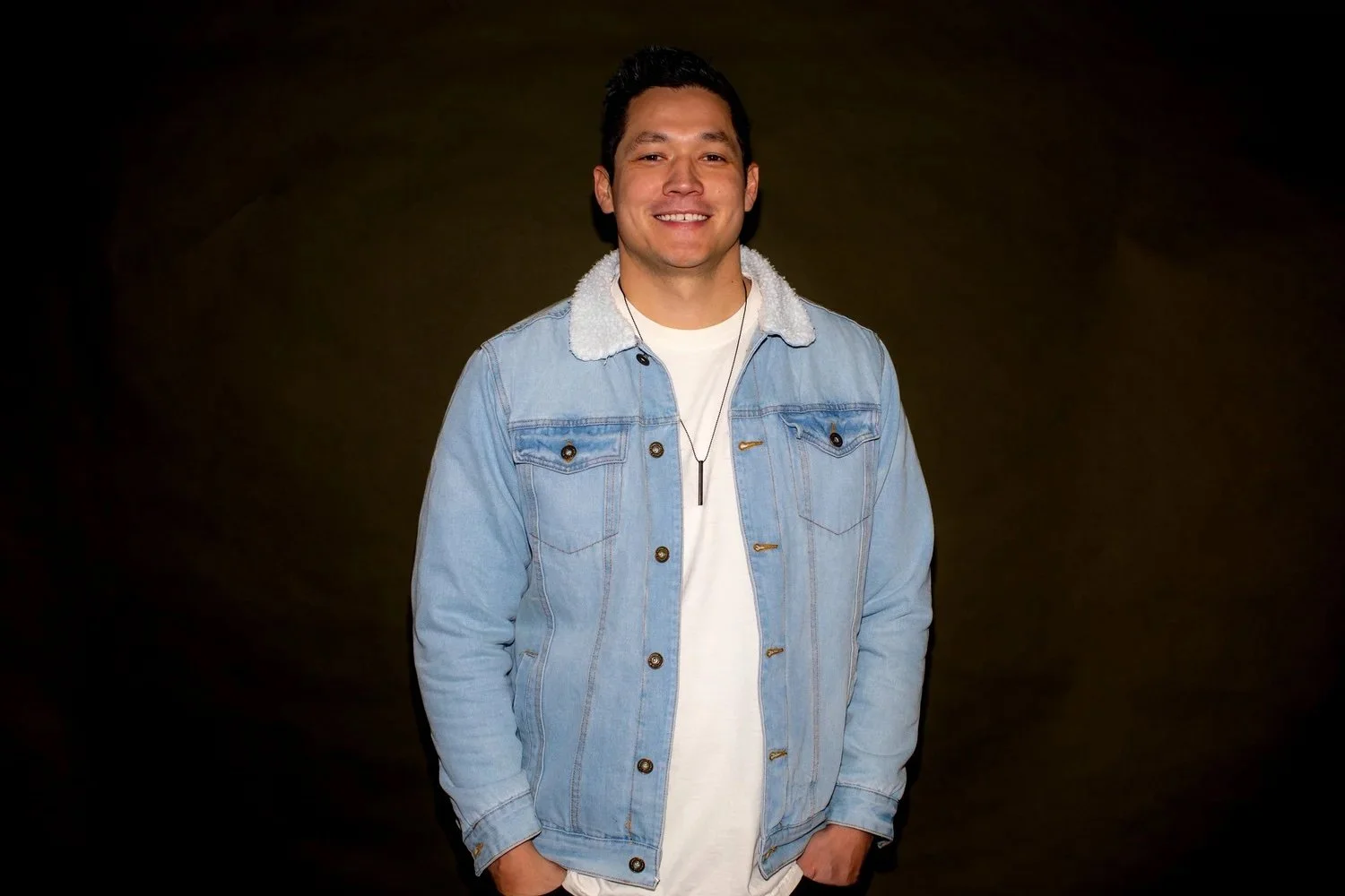 A young man with dark hair smiling, wearing a light blue denim jacket with a white fleece collar, a white shirt, and a black necklace, standing against a dark background.