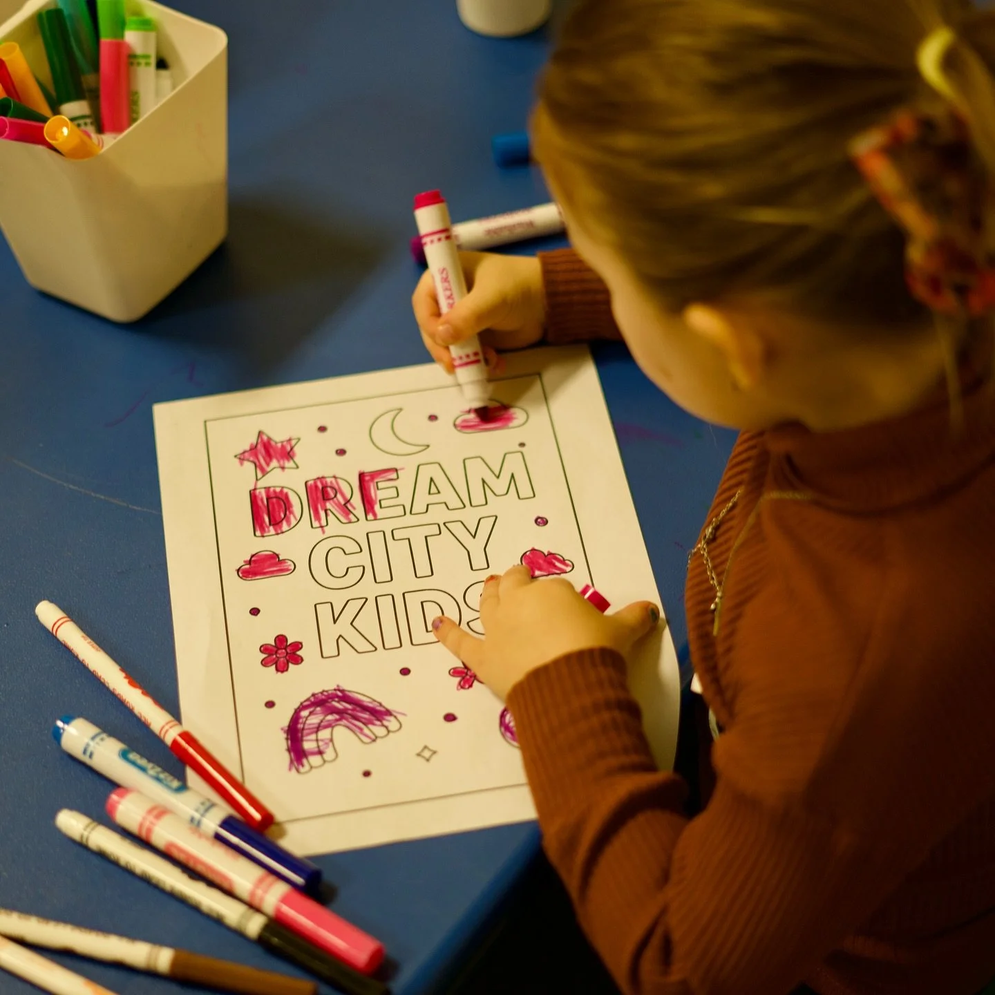 Child coloring a poster with the words 'DREAM CITY KIDS' using markers, surrounded by drawings of a rainbow, stars, moon, clouds, and various decorative elements on a blue table.