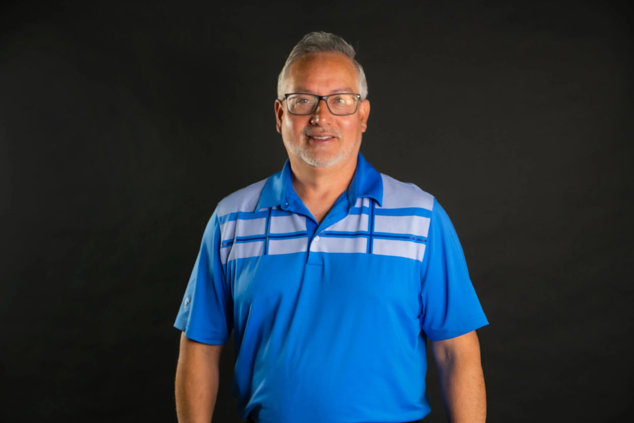A middle-aged man with gray hair, glasses, and a beard wearing a blue and gray polo shirt, standing against a dark background.